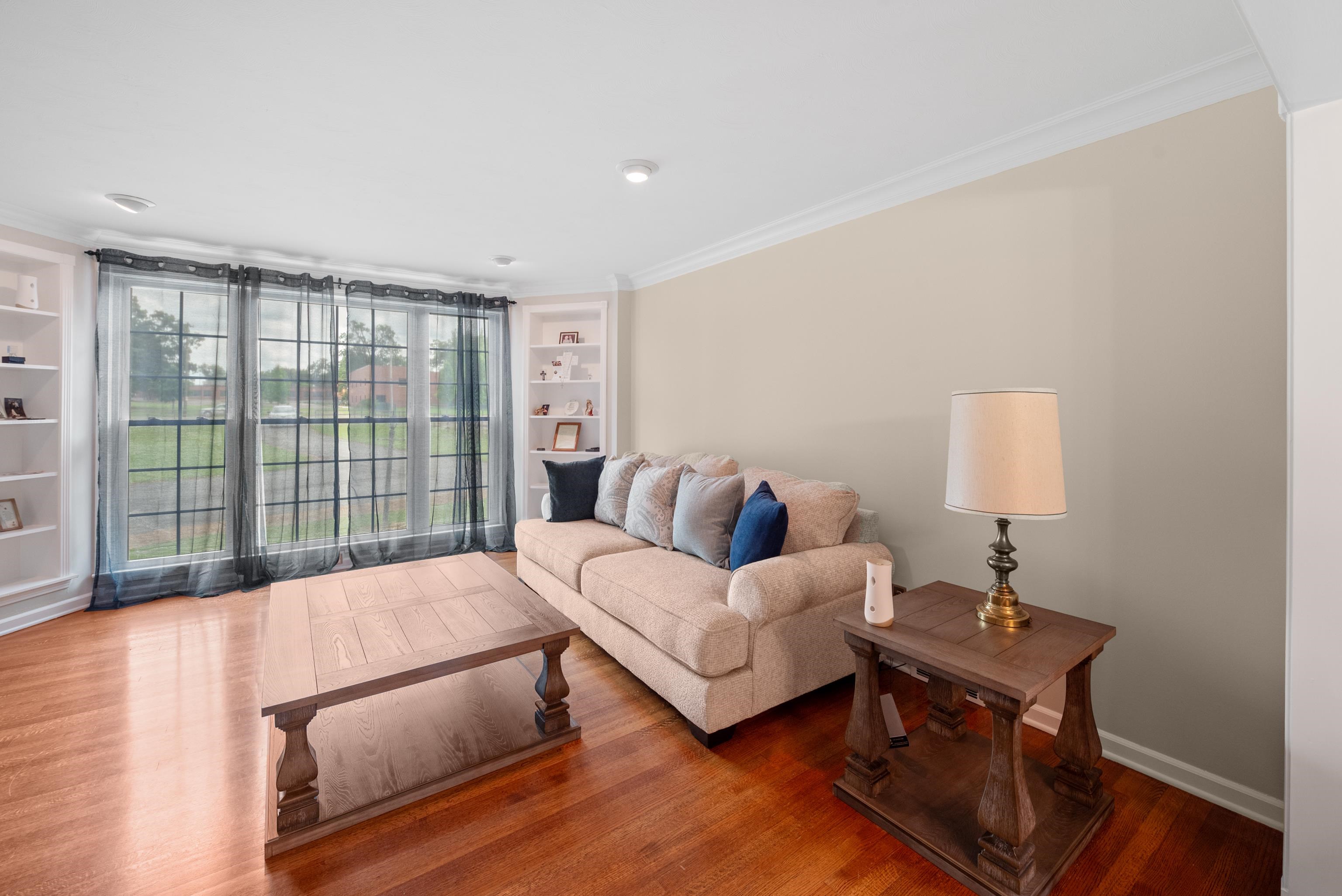 Living room with a beige sectional sofa, dark wood tables, hardwood floors, and large curtained windows.
