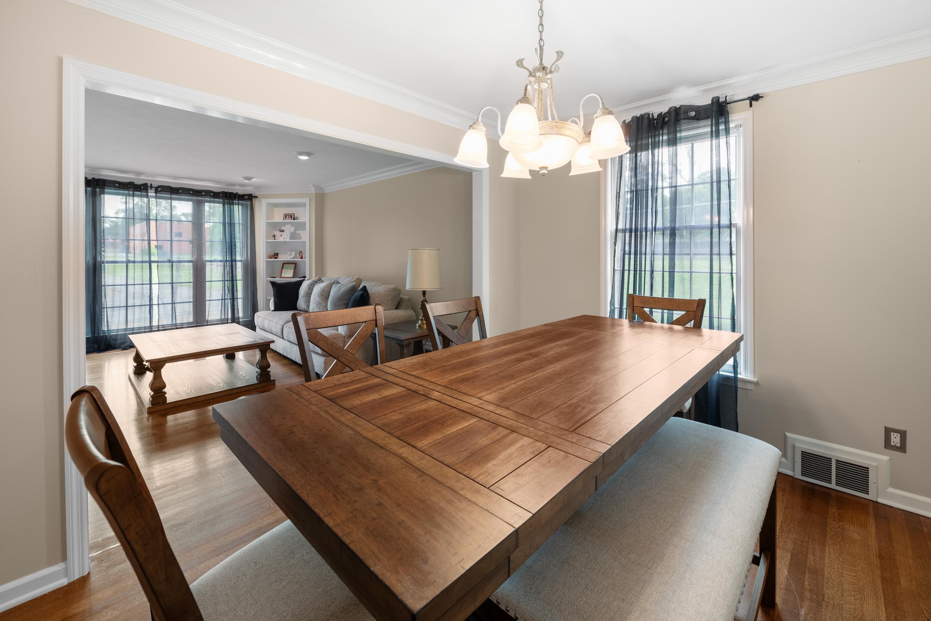 Dining room with a large wooden table, upholstered bench, chandelier, and adjoining living room.