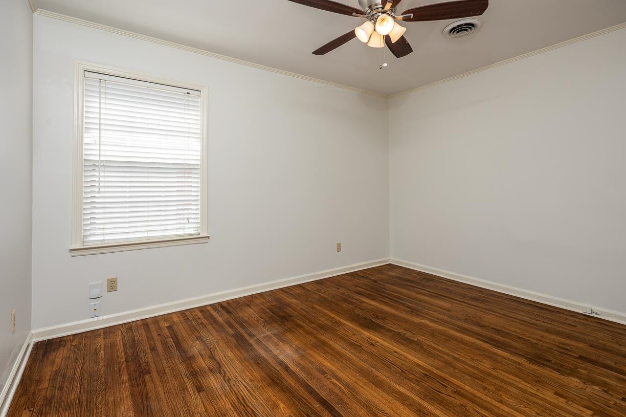 Empty bedroom with dark hardwood floors, white blinds, and a ceiling fan with lights.
