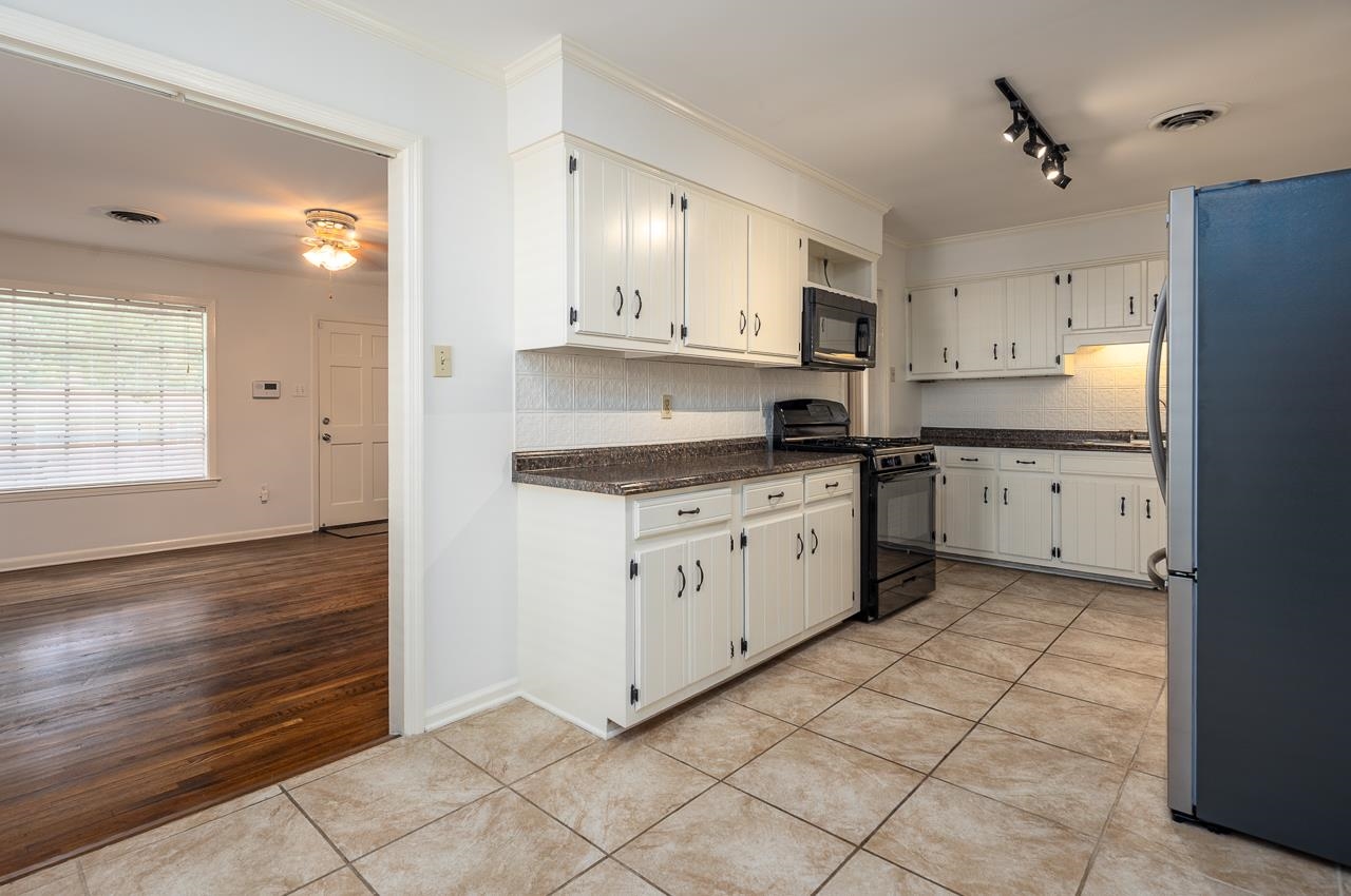 Kitchen with white cabinets, dark countertops, black appliances, and tan tile flooring opening to a wood-floored room.