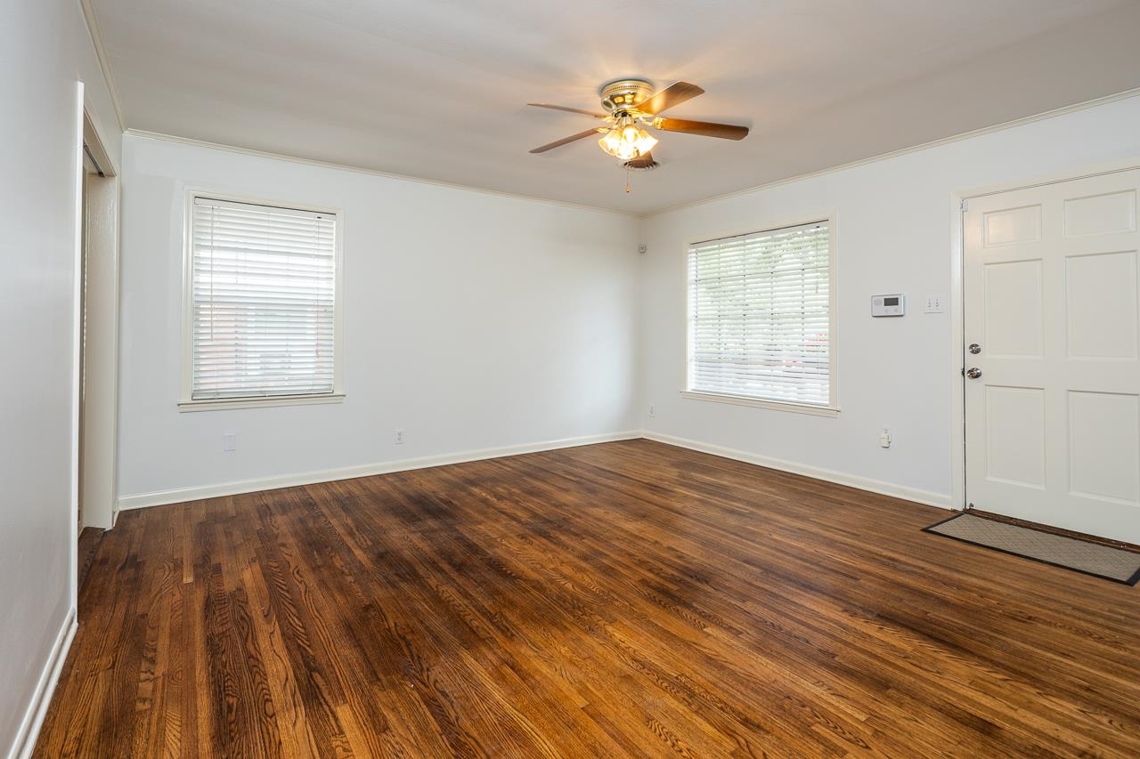 Empty room with dark hardwood floors, two blinded windows, and a ceiling fan with light.