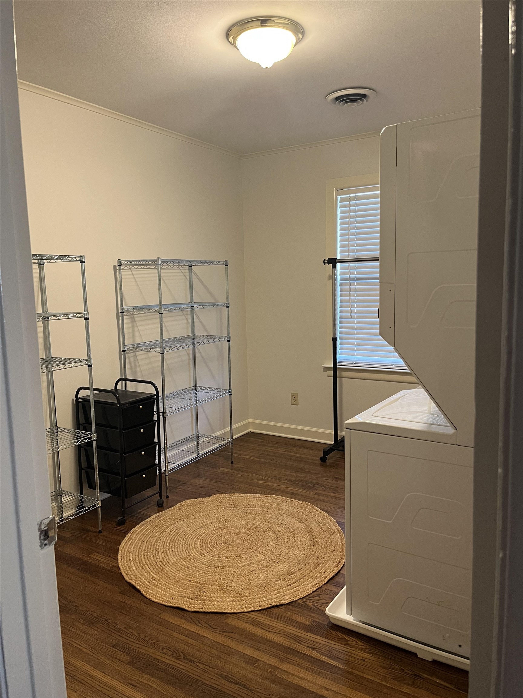 Laundry room with stacked appliances, wire shelving, and a round woven rug.