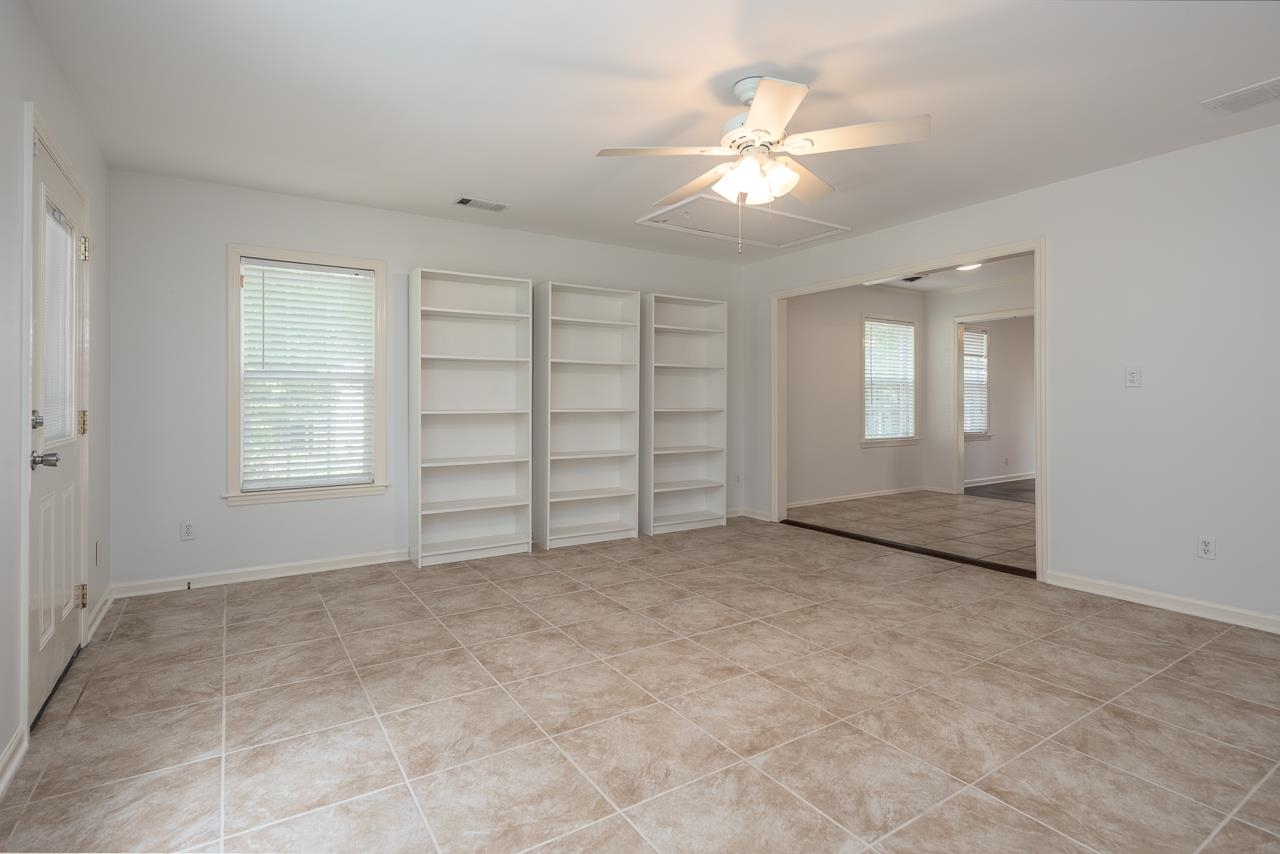 Empty tiled room with white bookshelves and a ceiling fan, opening into an adjoining room.