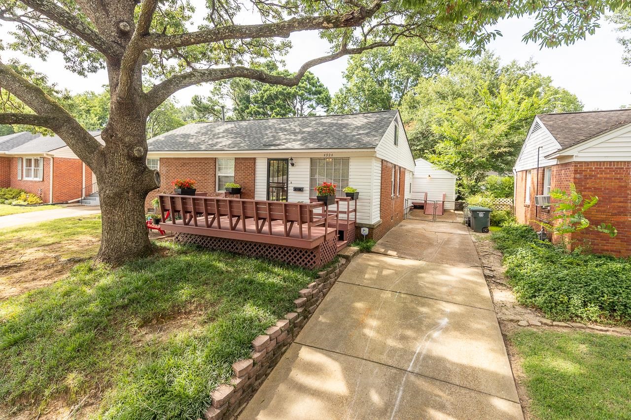 Single-story brick home with a raised deck and flower planters beneath a large shade tree.