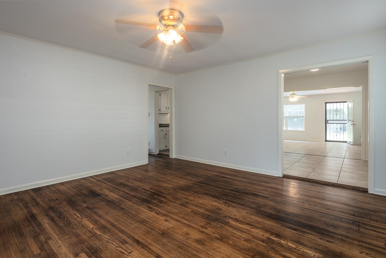 Empty living room with dark wood floors and ceiling fan, opening into a bright tiled room.