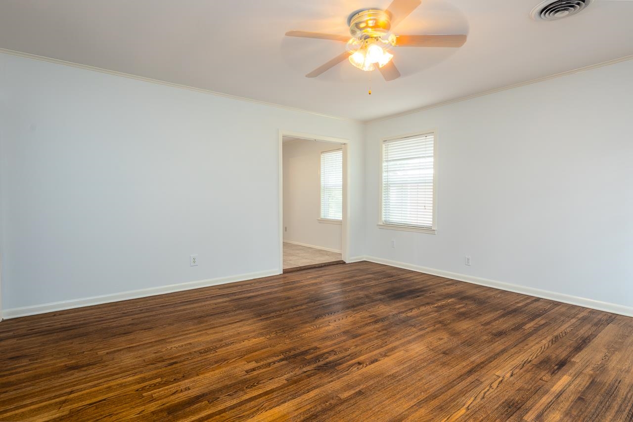 Empty room with dark hardwood floors, a lit ceiling fan, and windows with blinds.