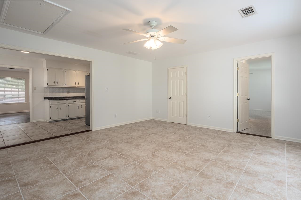Open tiled living area beside a white-cabinet kitchen, with a ceiling fan and doorway to a carpeted room.