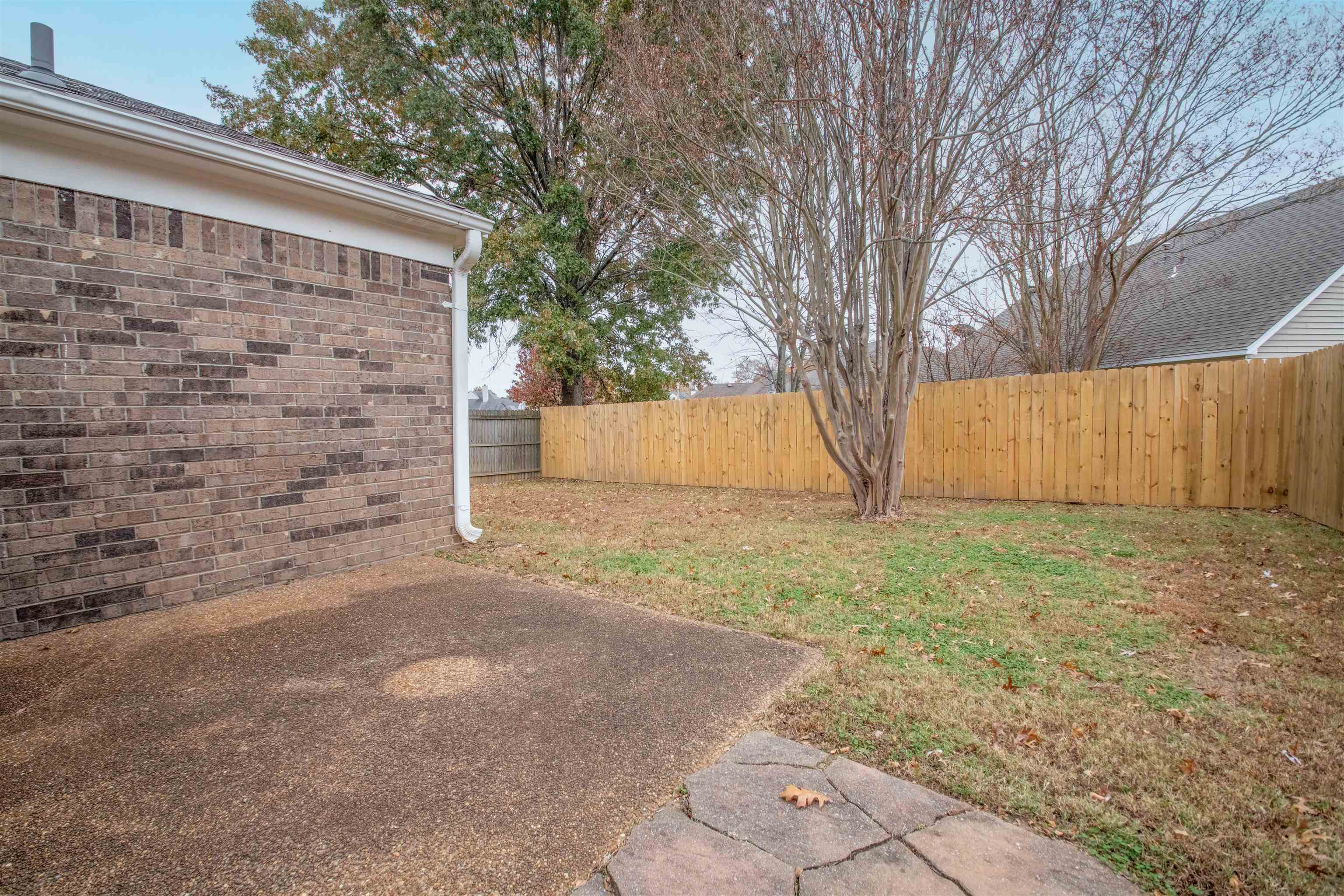 Backyard patio beside a brick house, with a wooden privacy fence and mature tree.