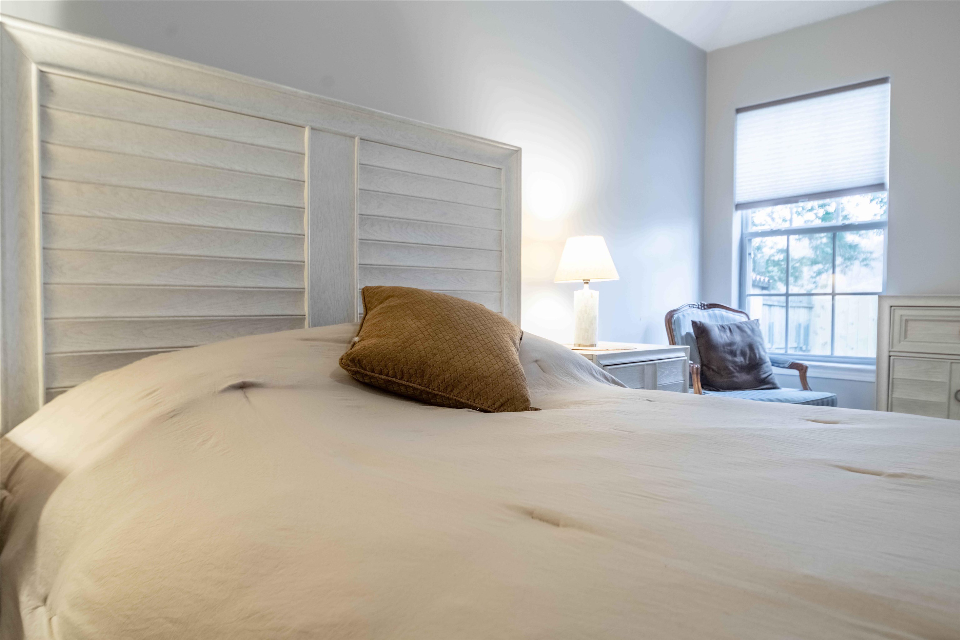 Staged bedroom with cream bedding, a paneled headboard, brown accent pillow, and window-side chair.