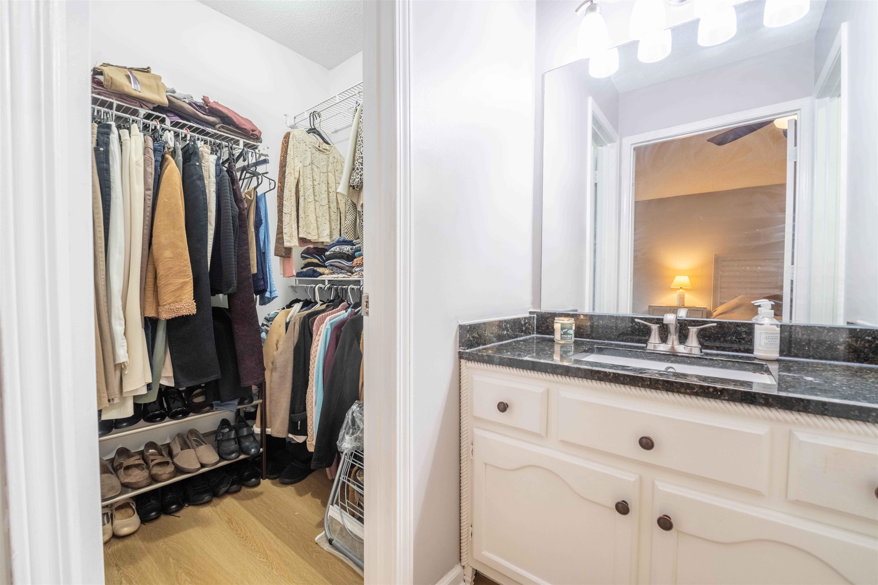 Walk-in closet filled with clothes and shoes beside a bathroom vanity with dark stone countertop.