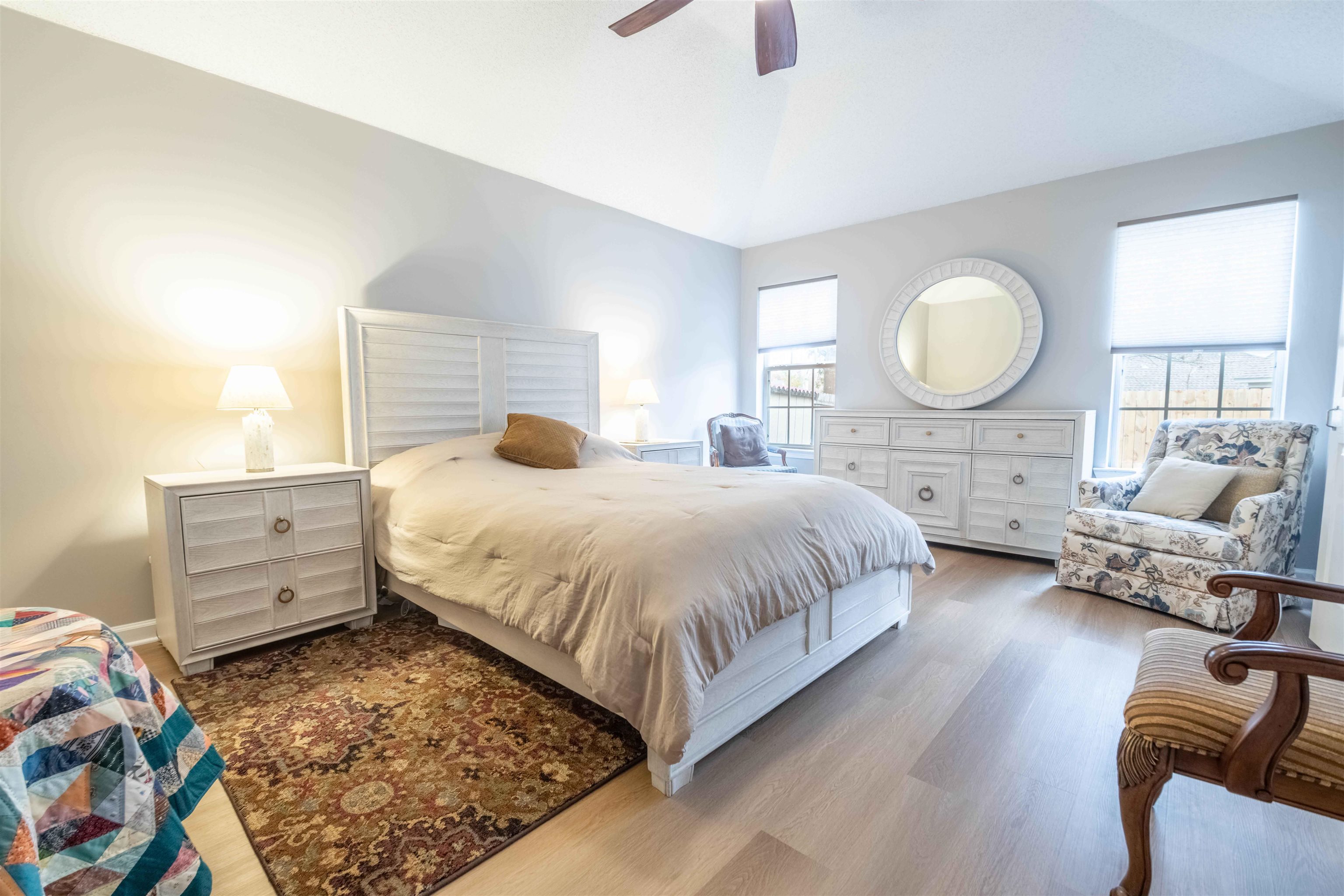 Bright bedroom with a large white bed, round mirror, floral armchair, and patterned rug.