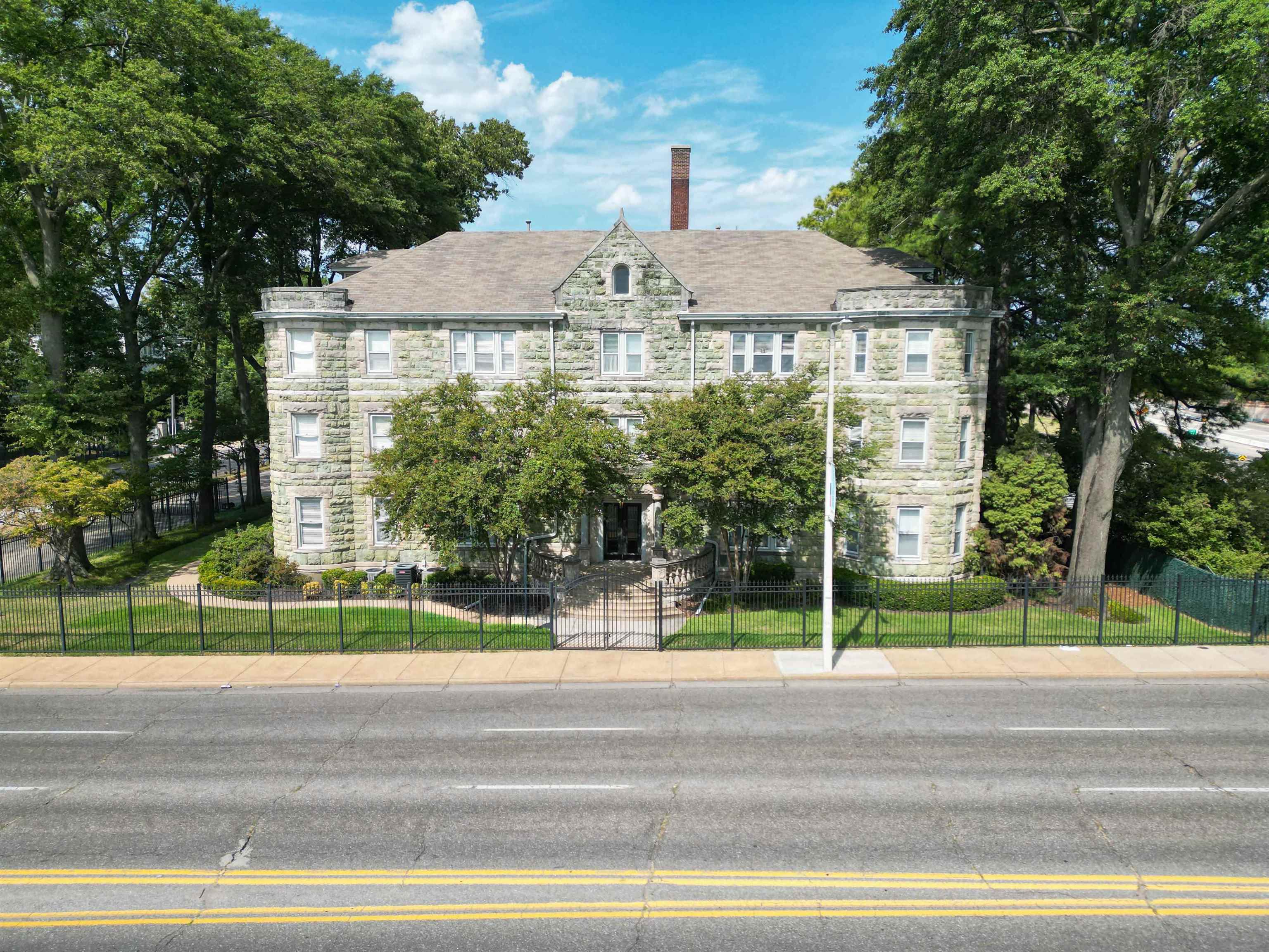 Stone apartment building with a fenced lawn, mature trees, and a central entrance.