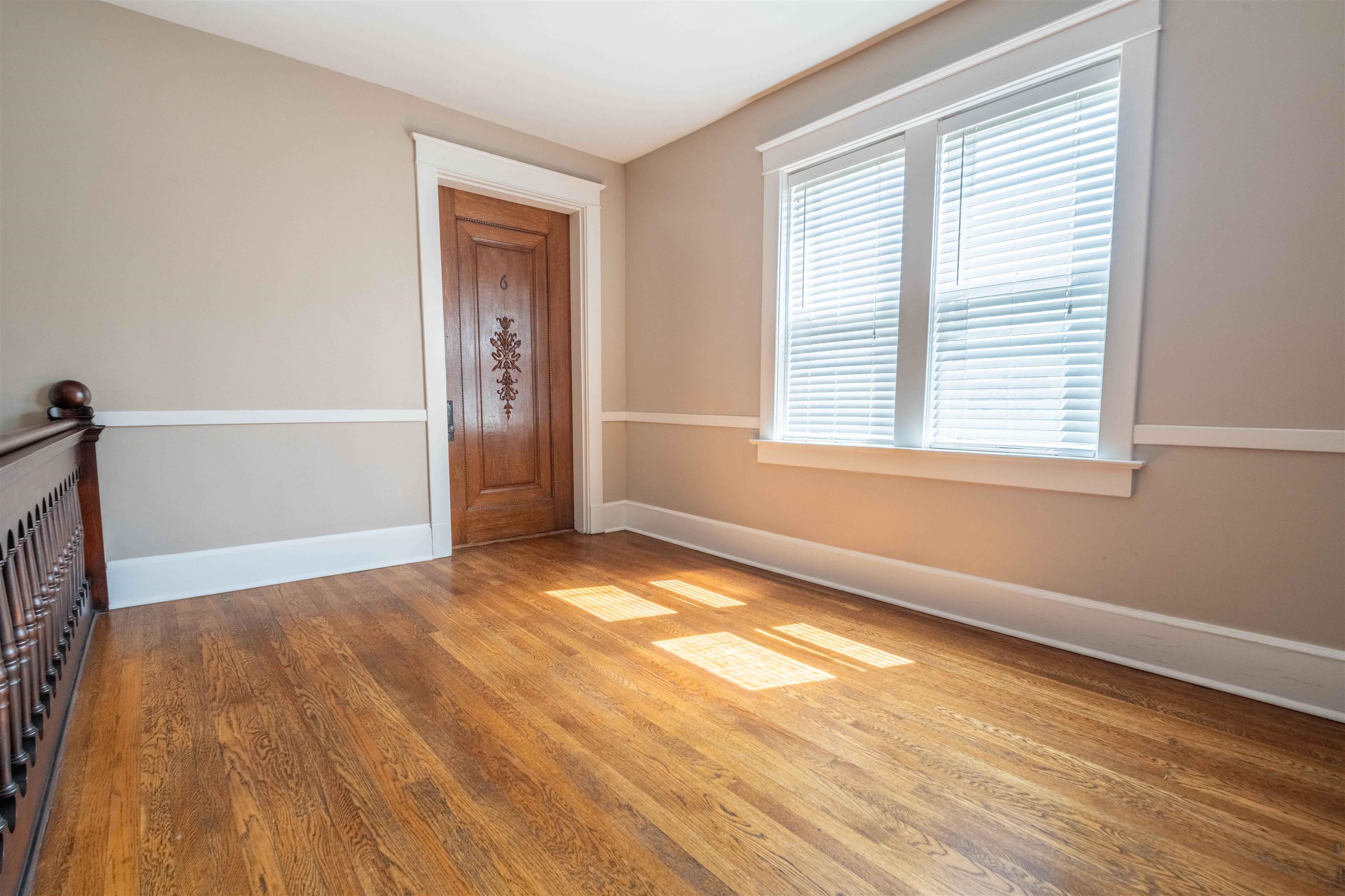 Empty room with hardwood floors, a carved wooden door, and sunlight streaming through a large window.