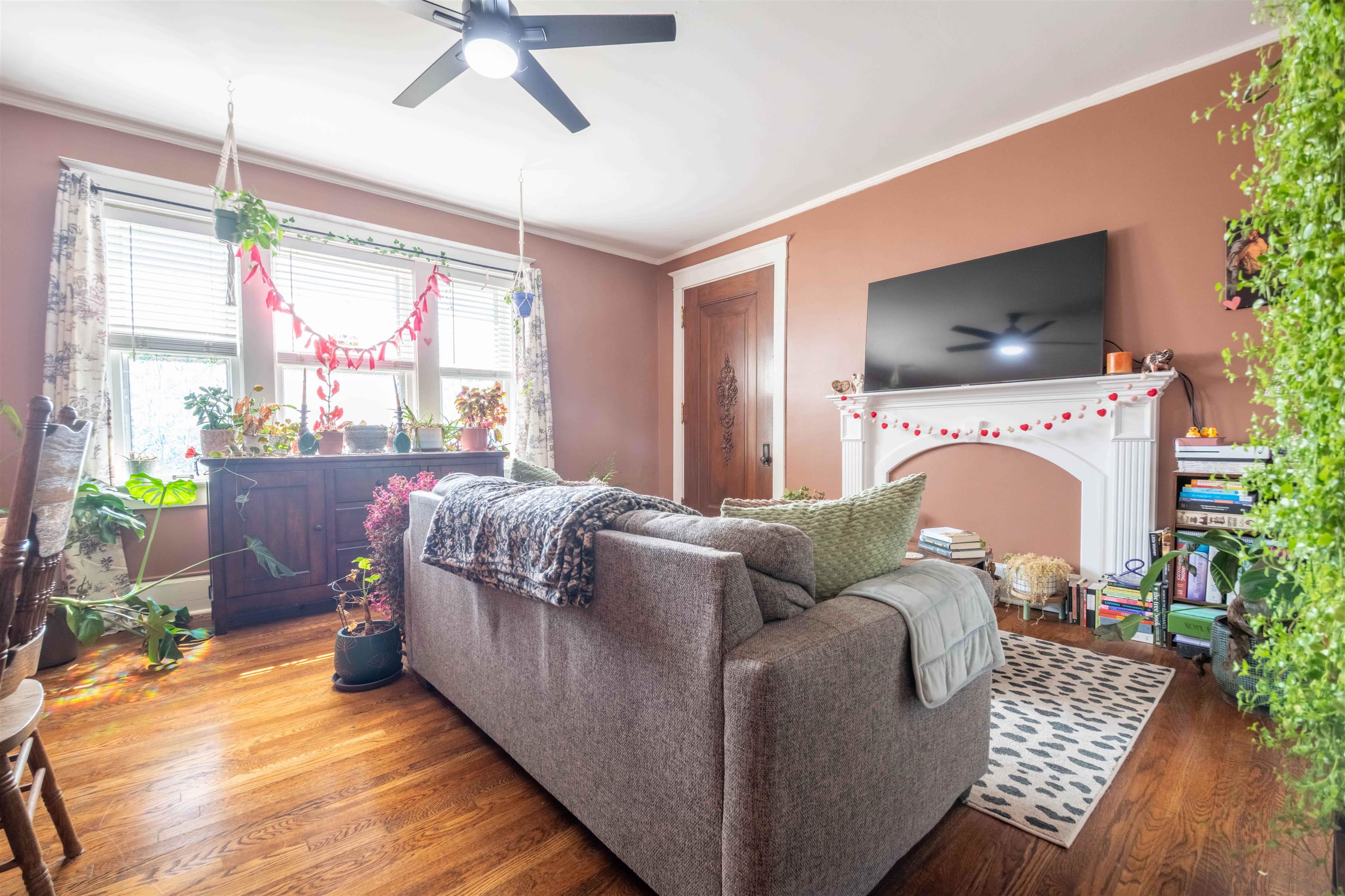 Living room with a gray sofa, hardwood floors, plant-filled windows, and a white fireplace.