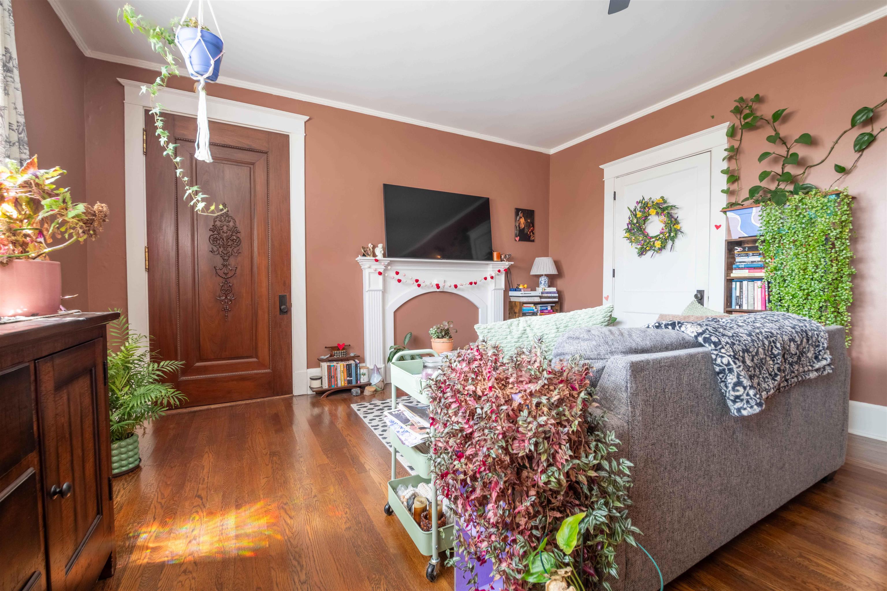 Living room with terracotta walls, hardwood floors, a decorative fireplace, gray sofa, and abundant plants.