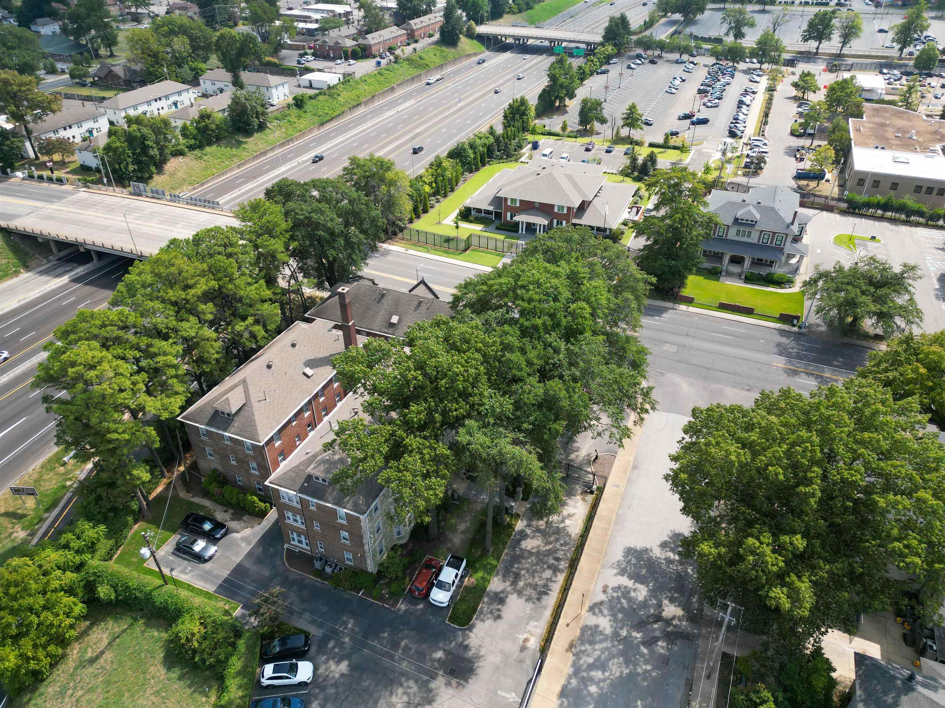 Aerial view of a brick apartment building surrounded by mature trees, parking and nearby roads.