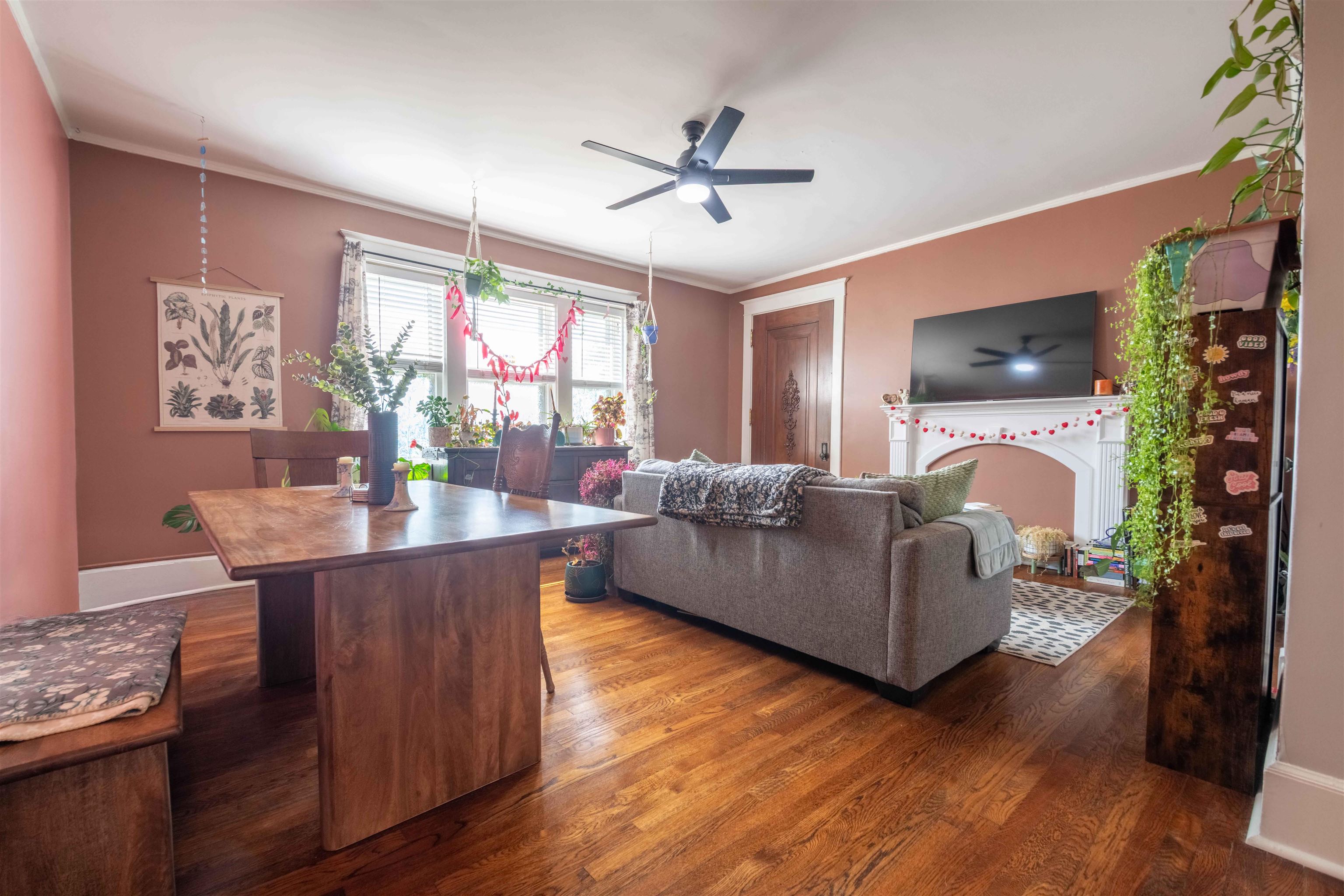 Furnished living and dining room with a gray sofa, white fireplace, hardwood floors, and abundant plants.