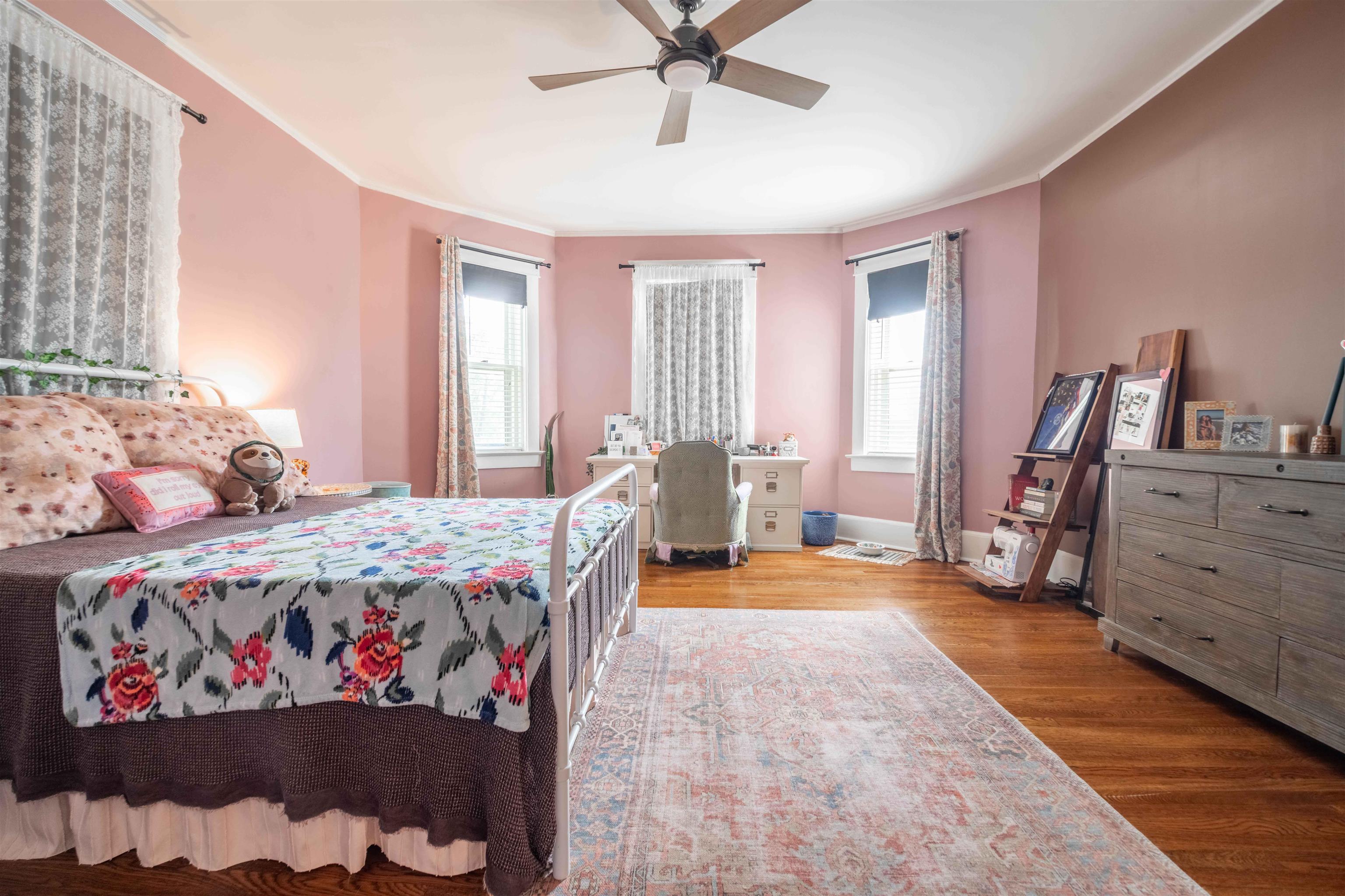 Bedroom with pink walls, floral bedding, hardwood floors, and a desk beneath three windows.