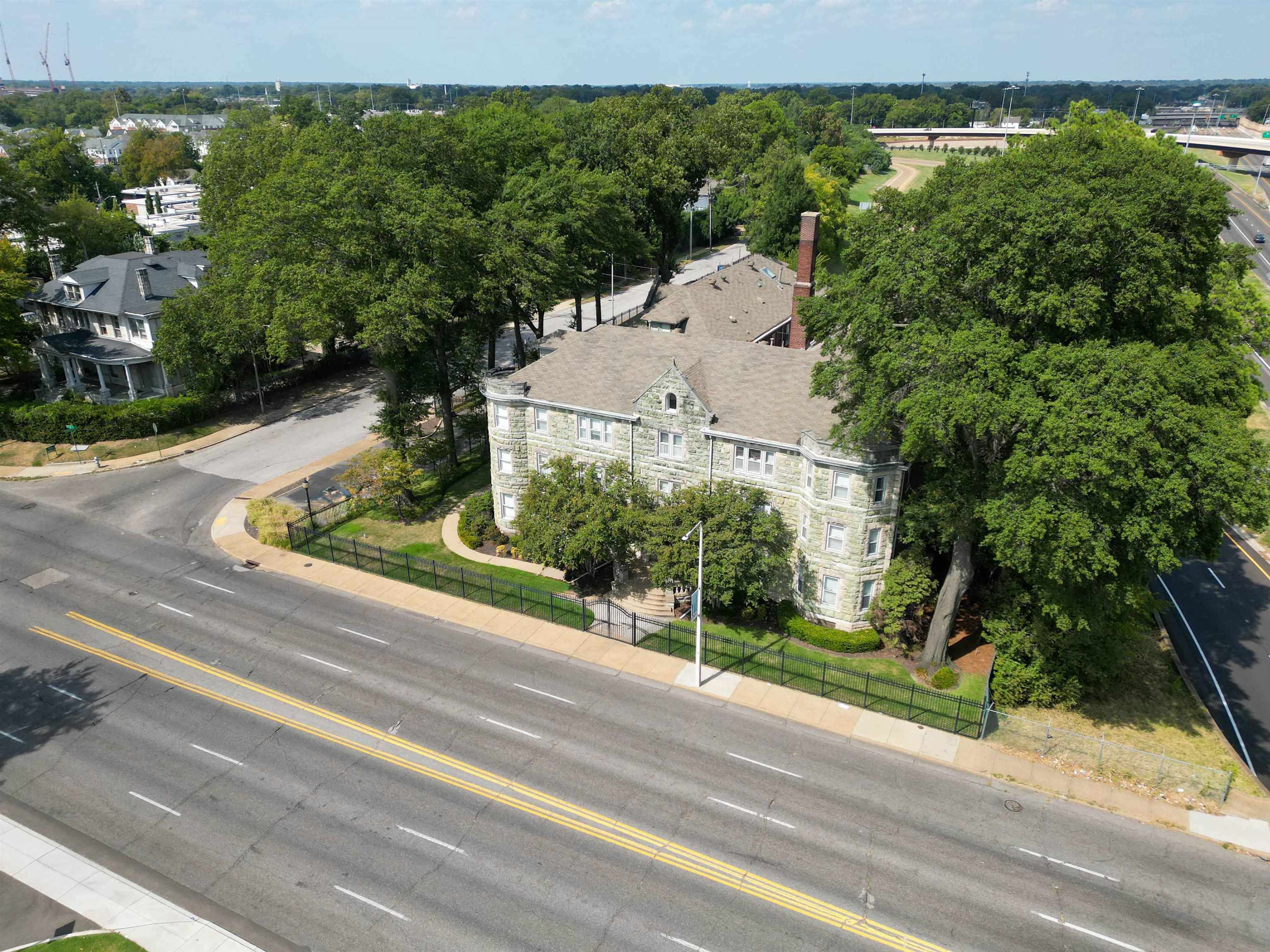 Aerial view of a stone residential building with a fenced lawn and mature trees beside a street.