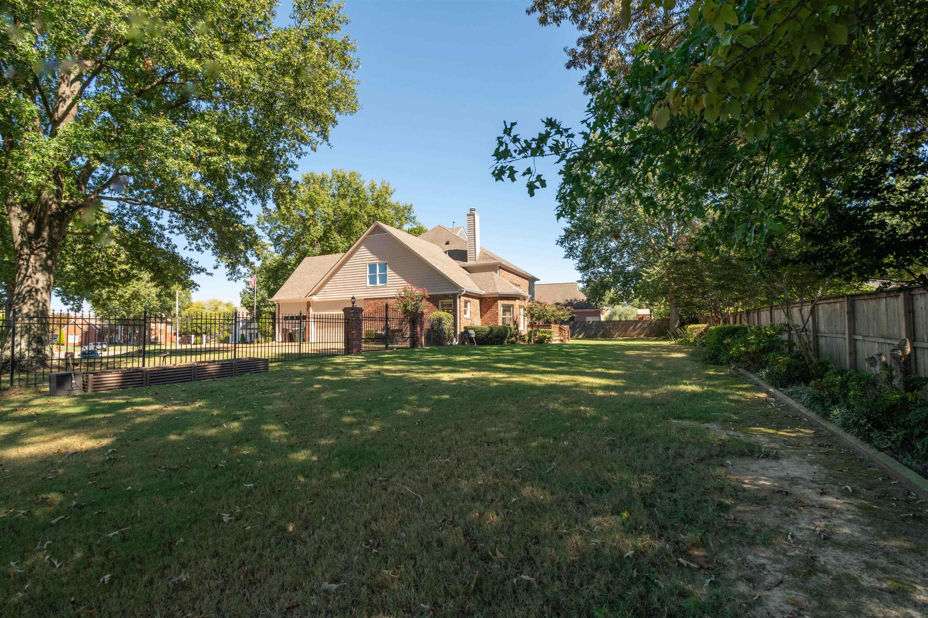 Rear of a brick-and-siding house beyond a shaded lawn, enclosed by fencing and mature trees.