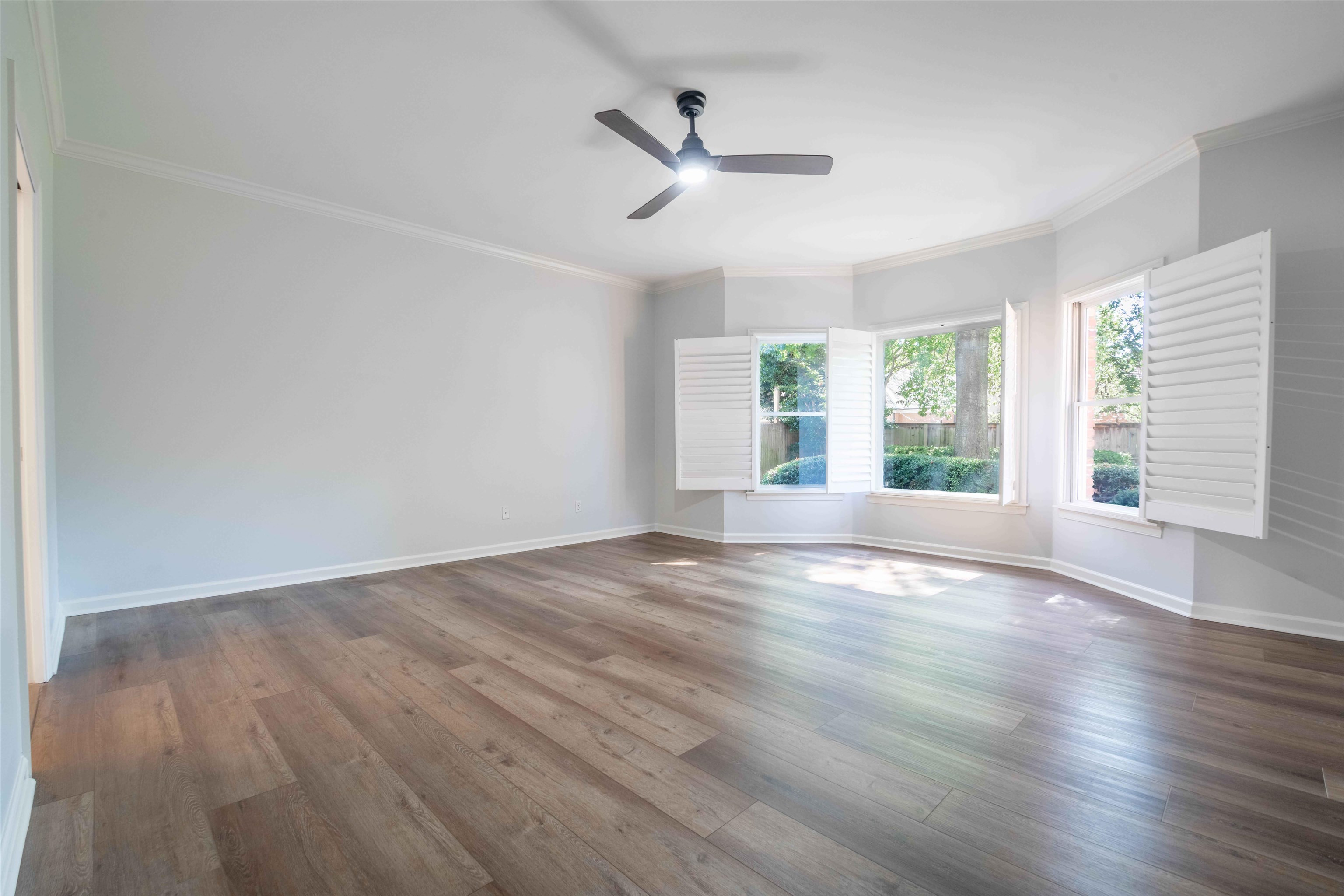 Empty room with wide-plank wood flooring, a ceiling fan, and windows with white shutters.