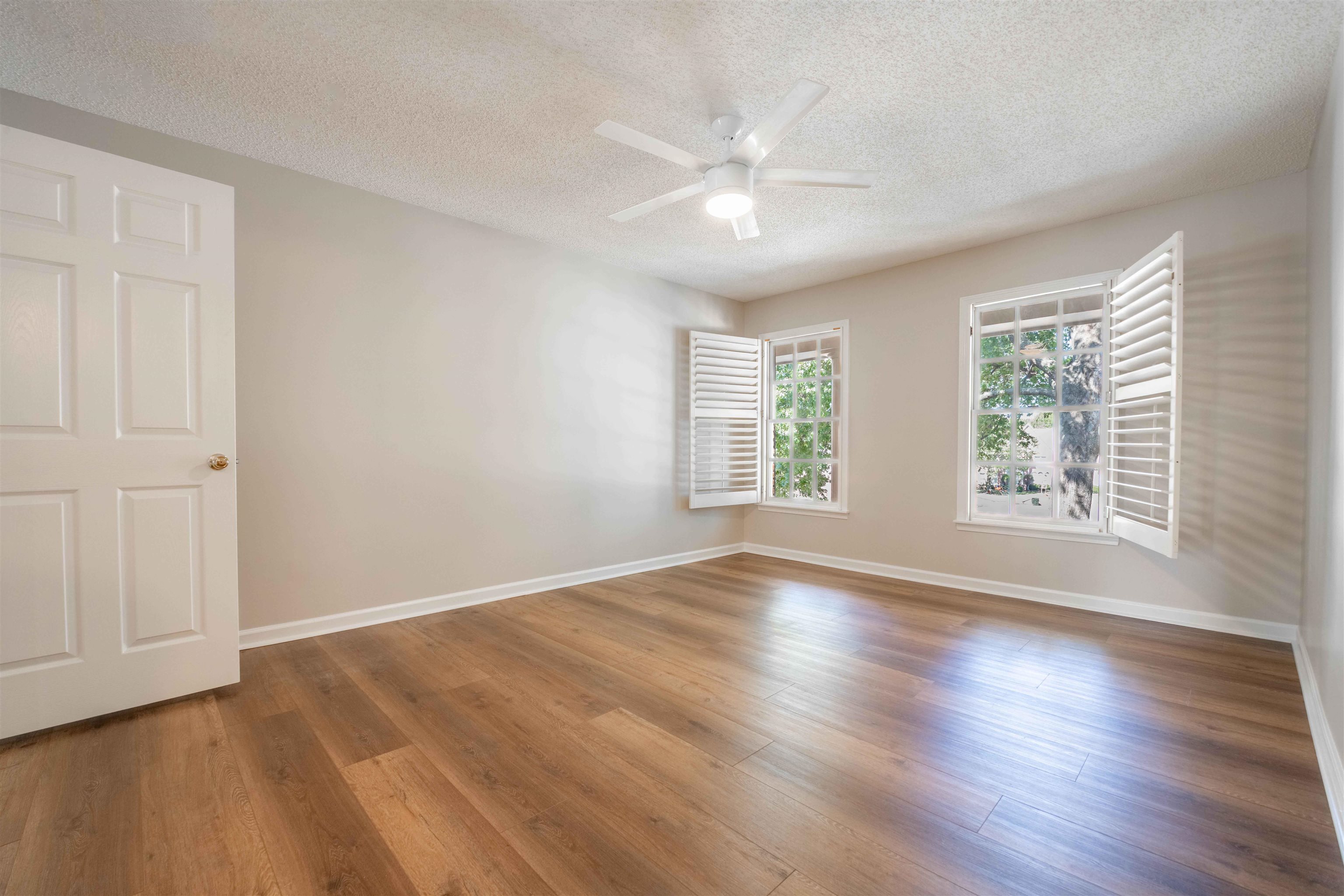 Empty bedroom with wood floors, shuttered windows, and a white ceiling fan.