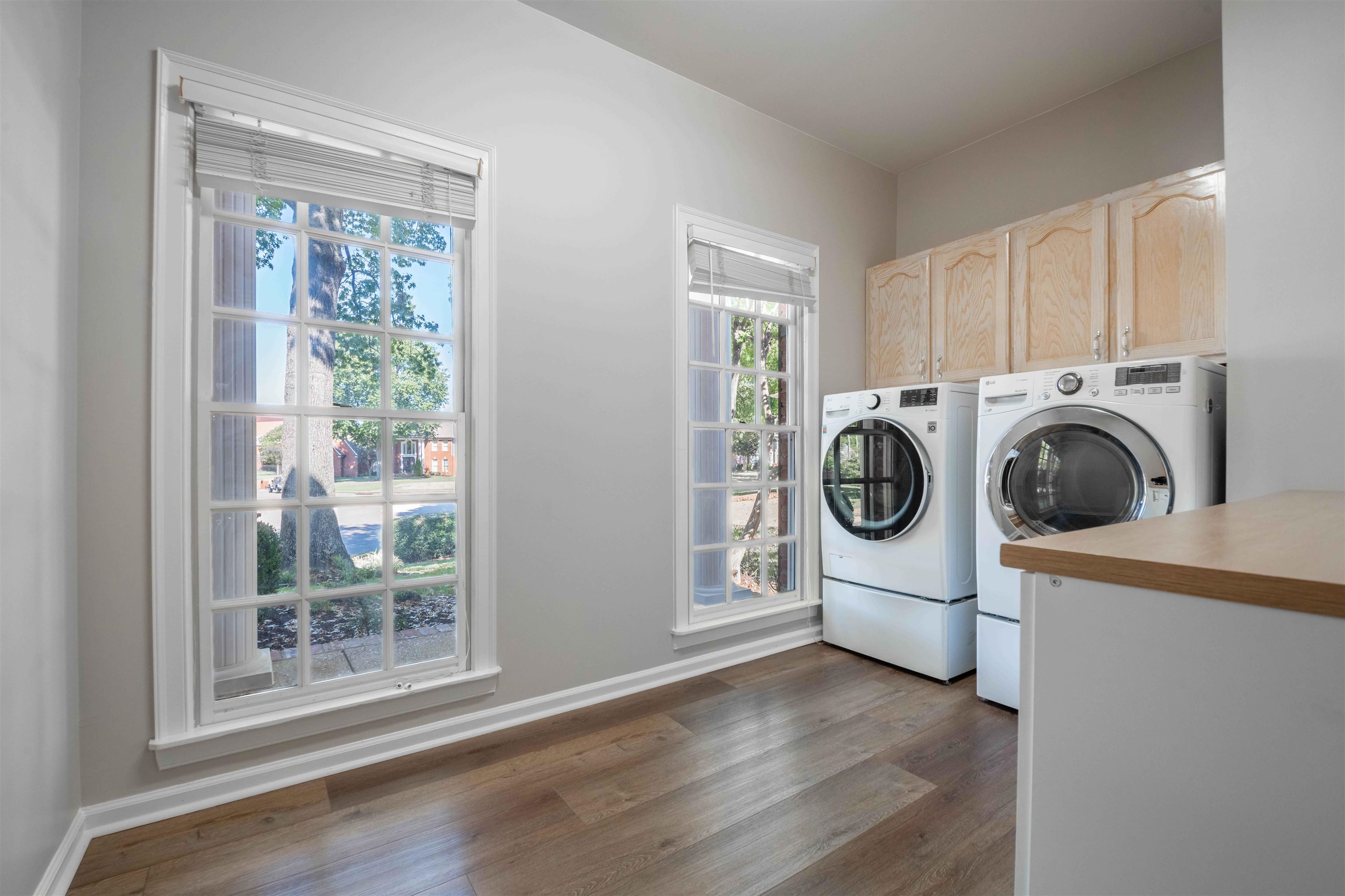 Laundry room with front-loading washer and dryer beneath wood cabinets, beside two large windows.