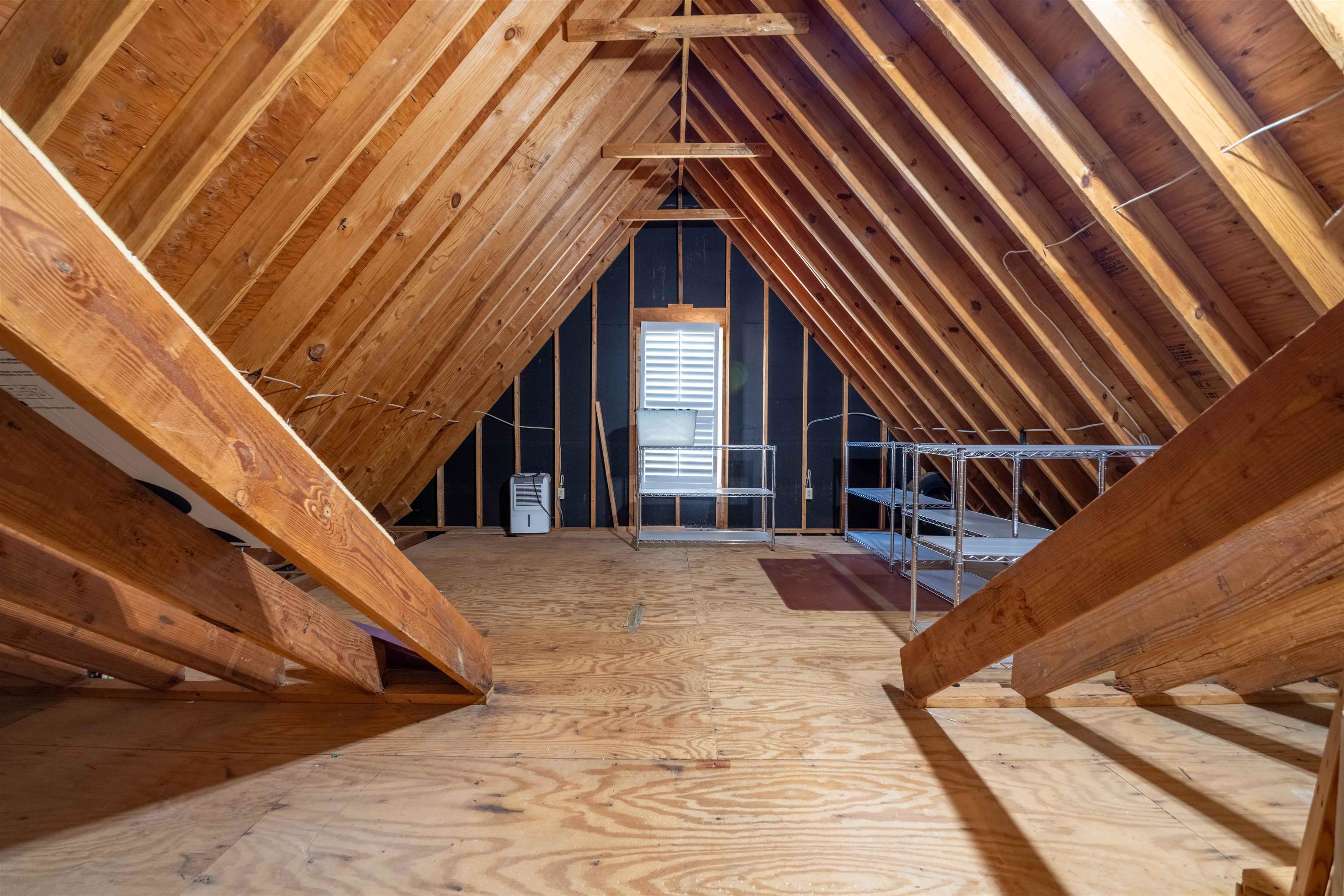 Unfinished attic with exposed wooden rafters, plywood flooring, metal shelving, and a shuttered window.
