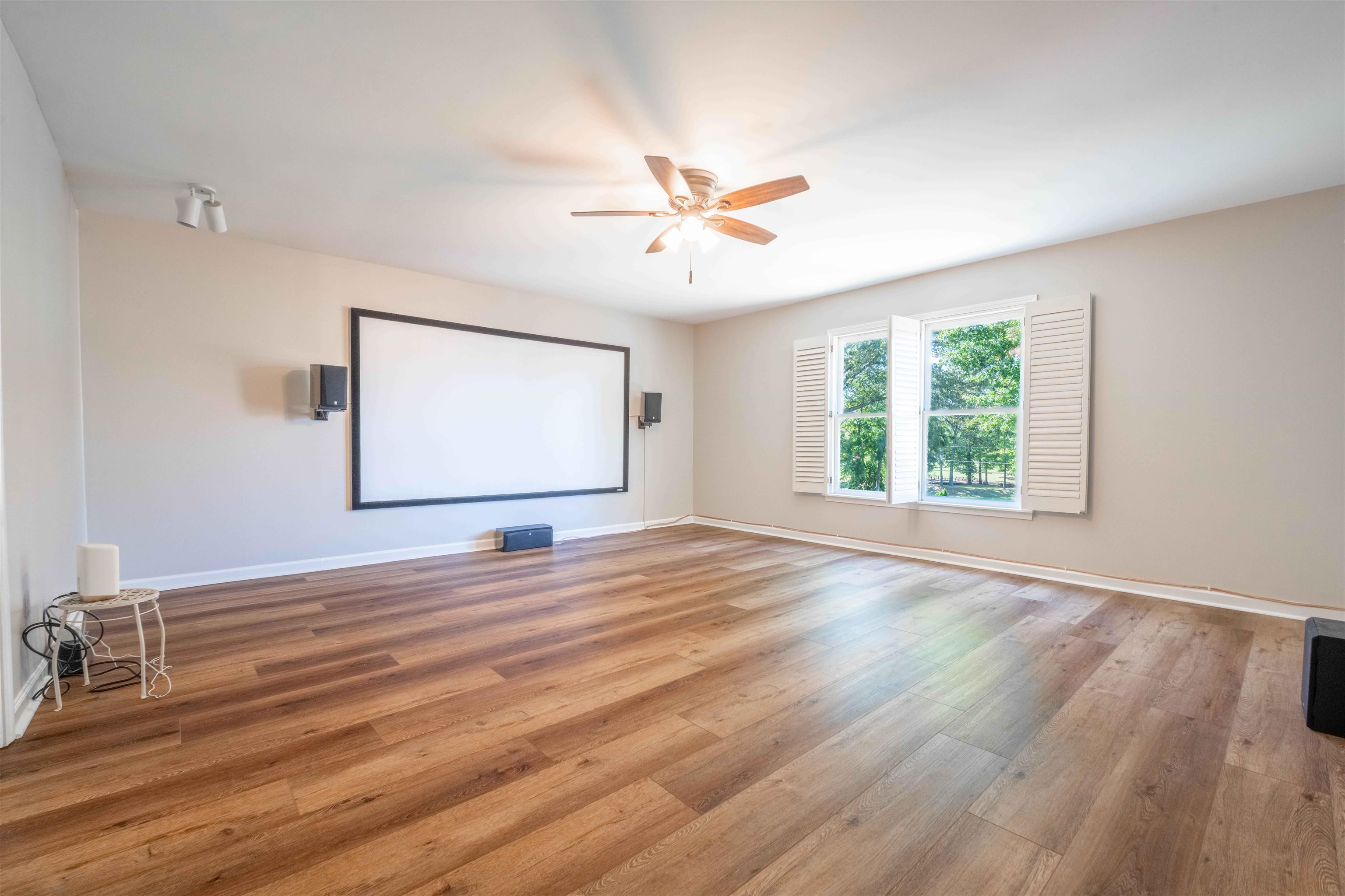 Empty media room with a large projection screen, wood flooring, and shuttered windows overlooking trees.
