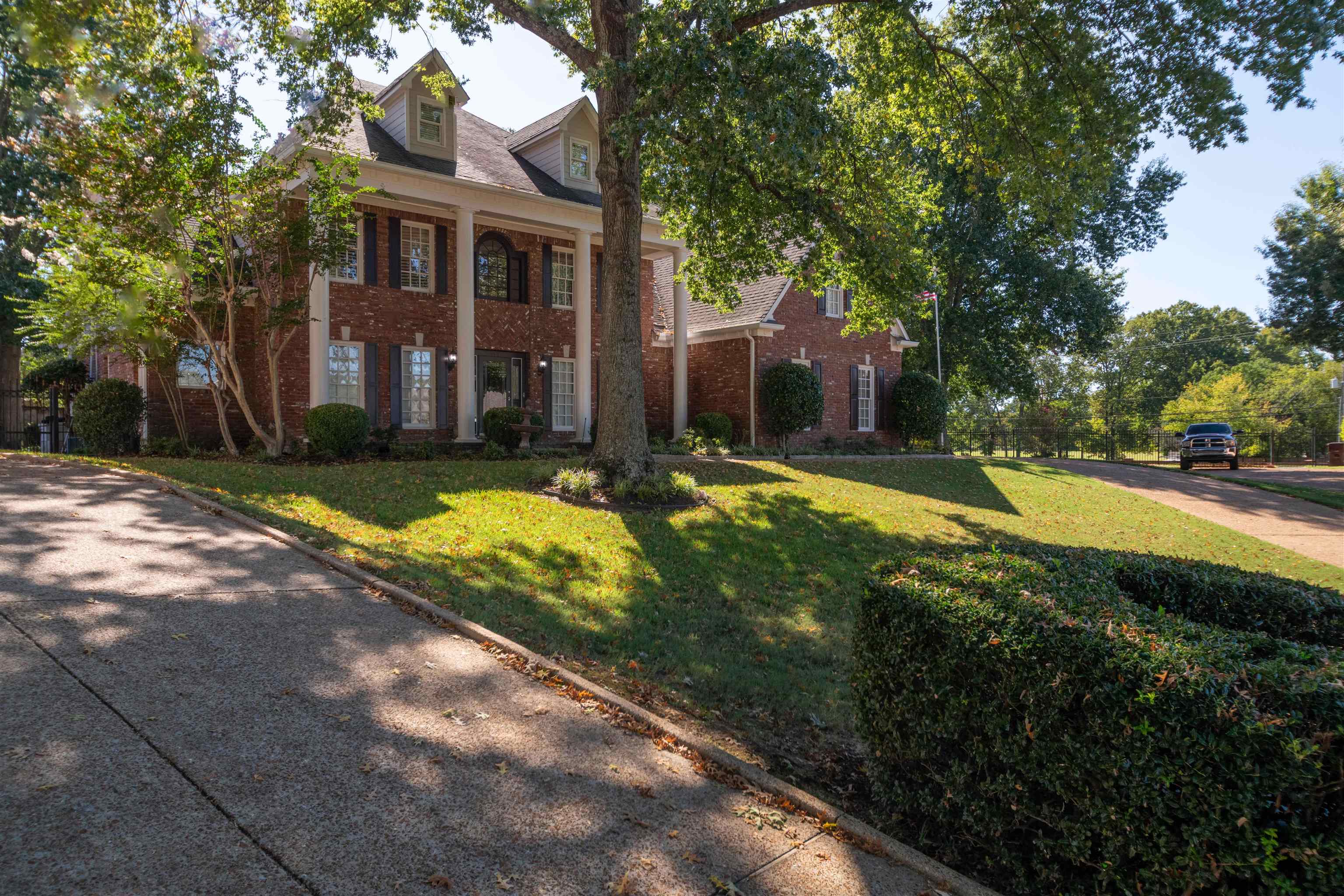 Brick two-story home with white columns, dormer windows, and mature trees shading the lawn.