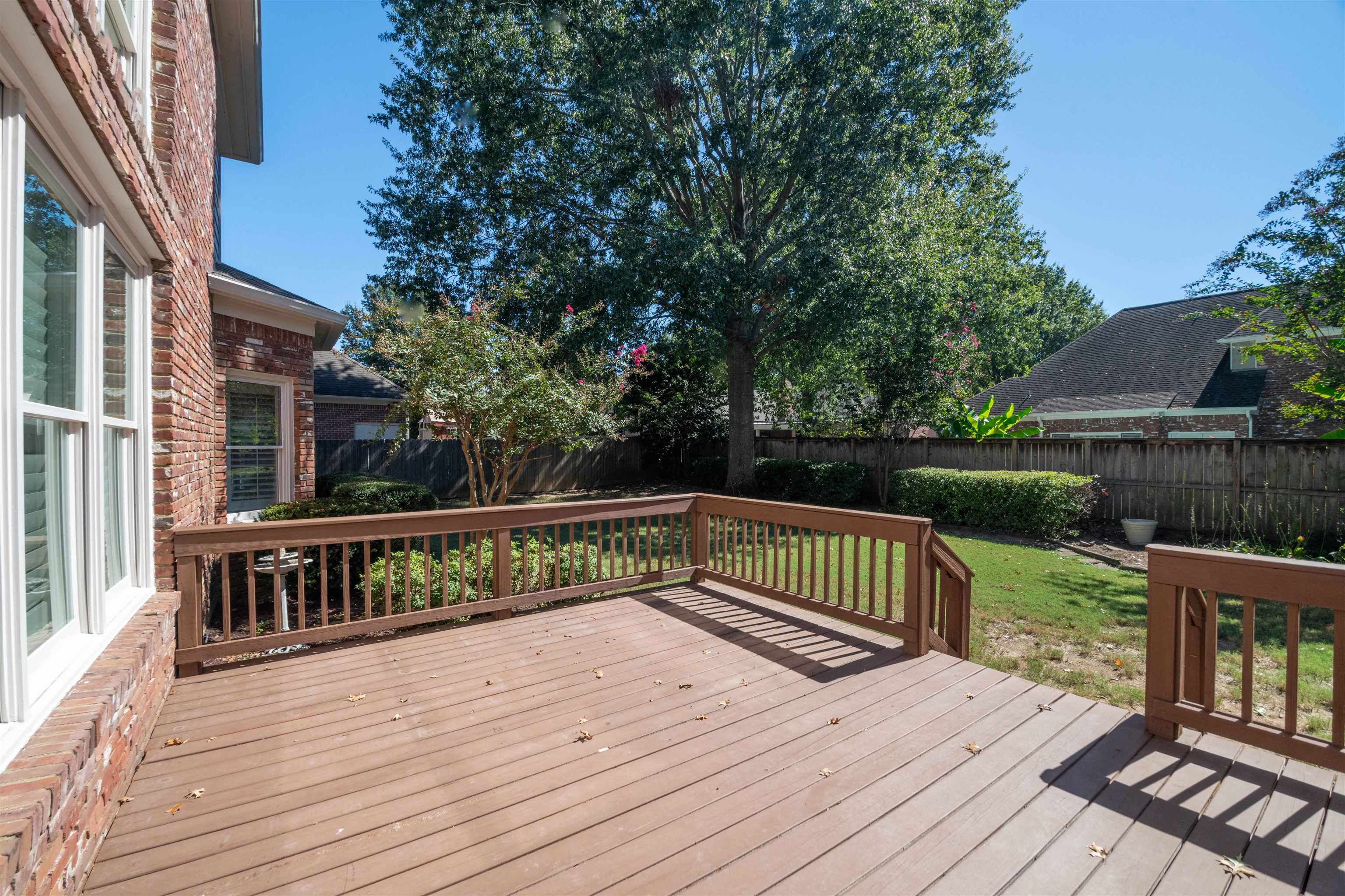 Backyard deck beside a brick house, overlooking a fenced lawn and mature trees.