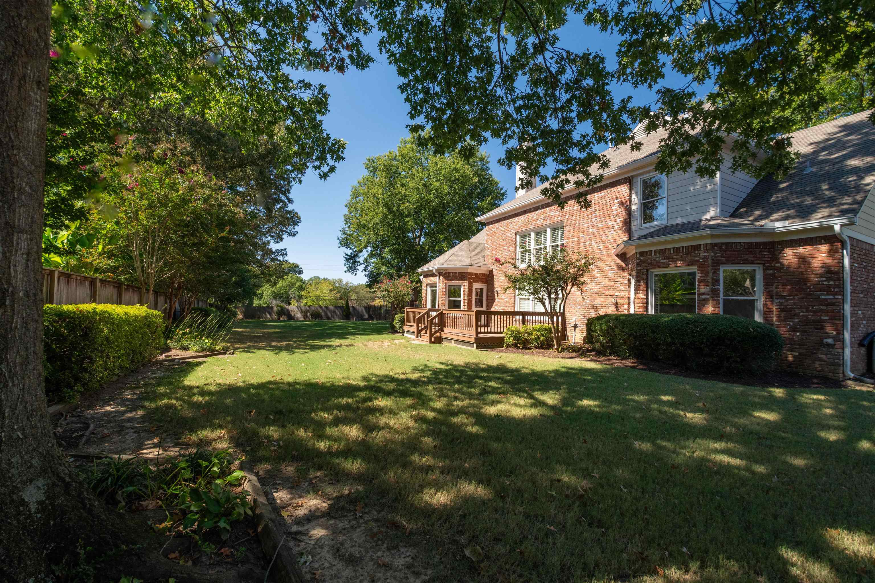 Brick home’s rear exterior with a wooden deck, broad lawn, and mature trees.