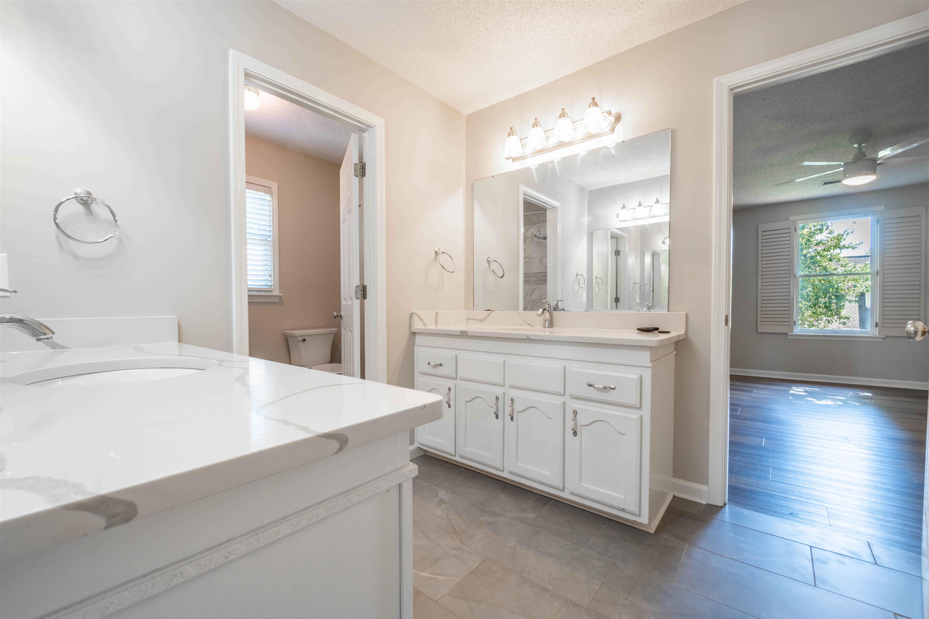 Bathroom with white double vanities and large mirrors, opening to a bedroom with a shuttered window.