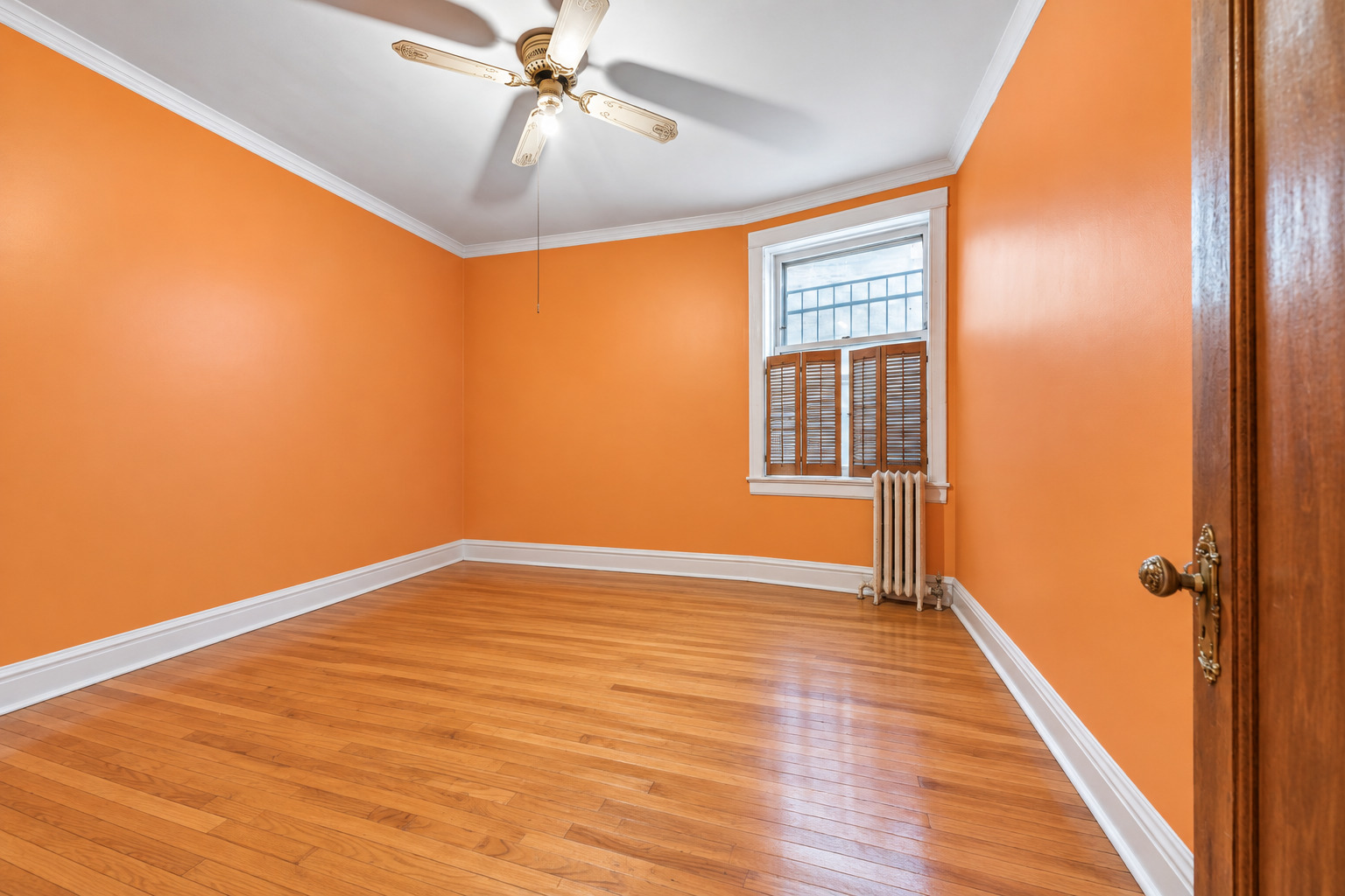 Empty orange-walled room with hardwood floors, a ceiling fan, and a shuttered window above a radiator.