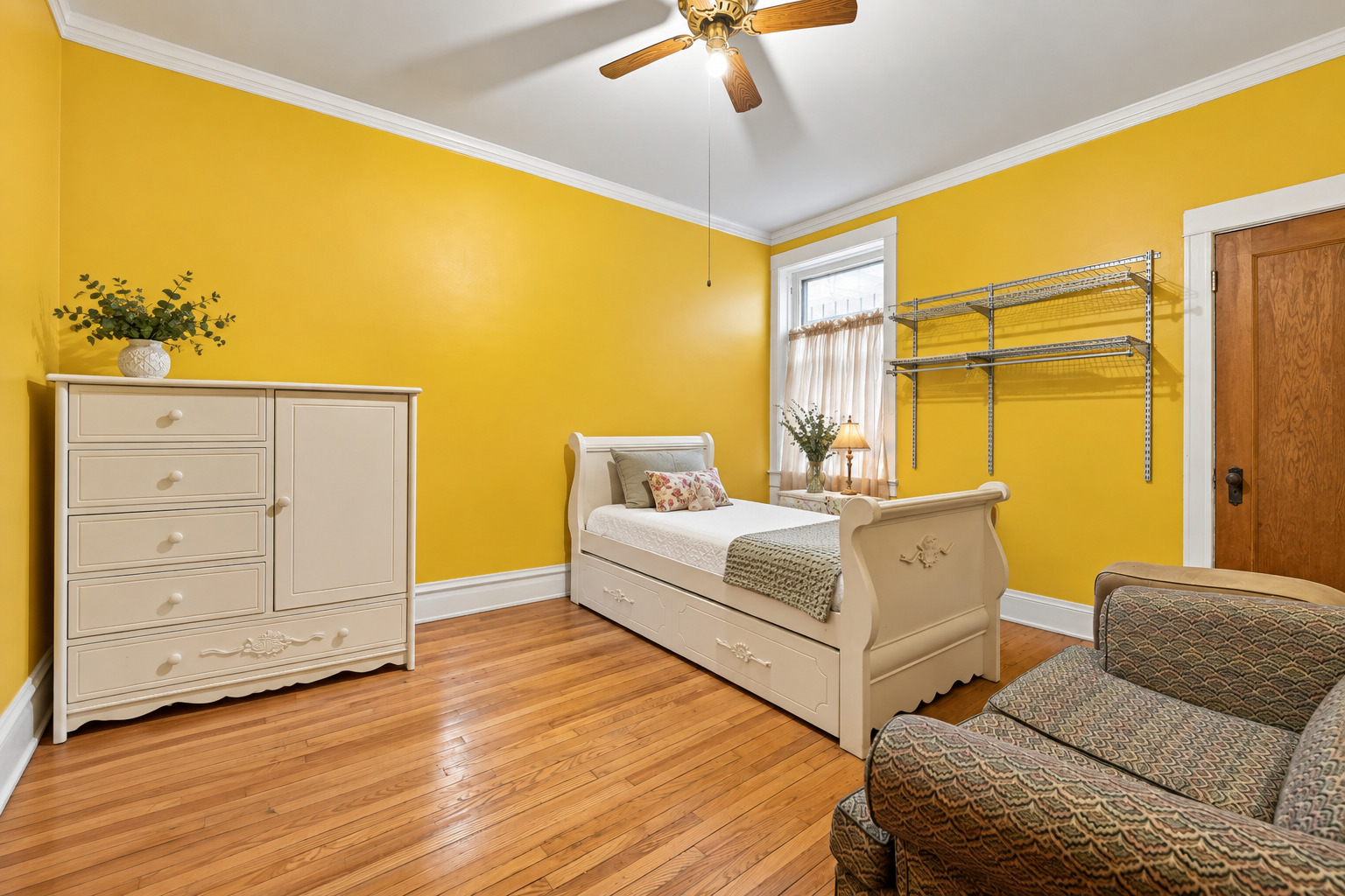 Bedroom with bright yellow walls, hardwood floors, a white bed, and a patterned chair.