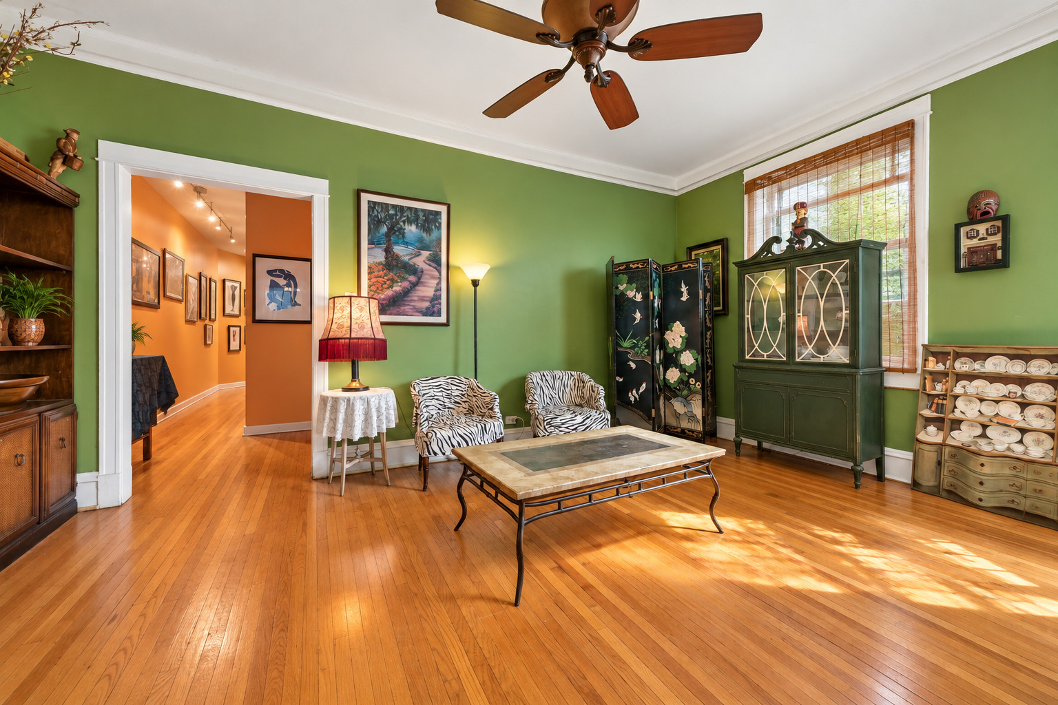 Living room with green walls, zebra-patterned chairs, hardwood floors, and sunlight streaming through bamboo blinds.