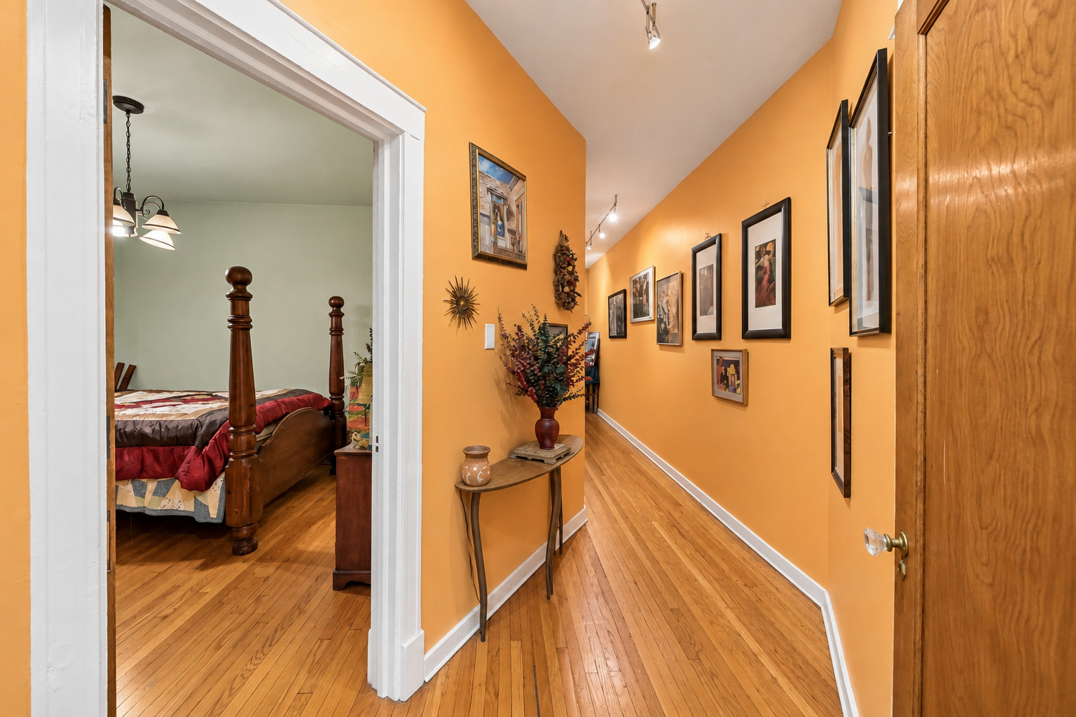 Orange-walled apartment hallway with hardwood floors and framed art, opening onto a bedroom with a four-poster bed.