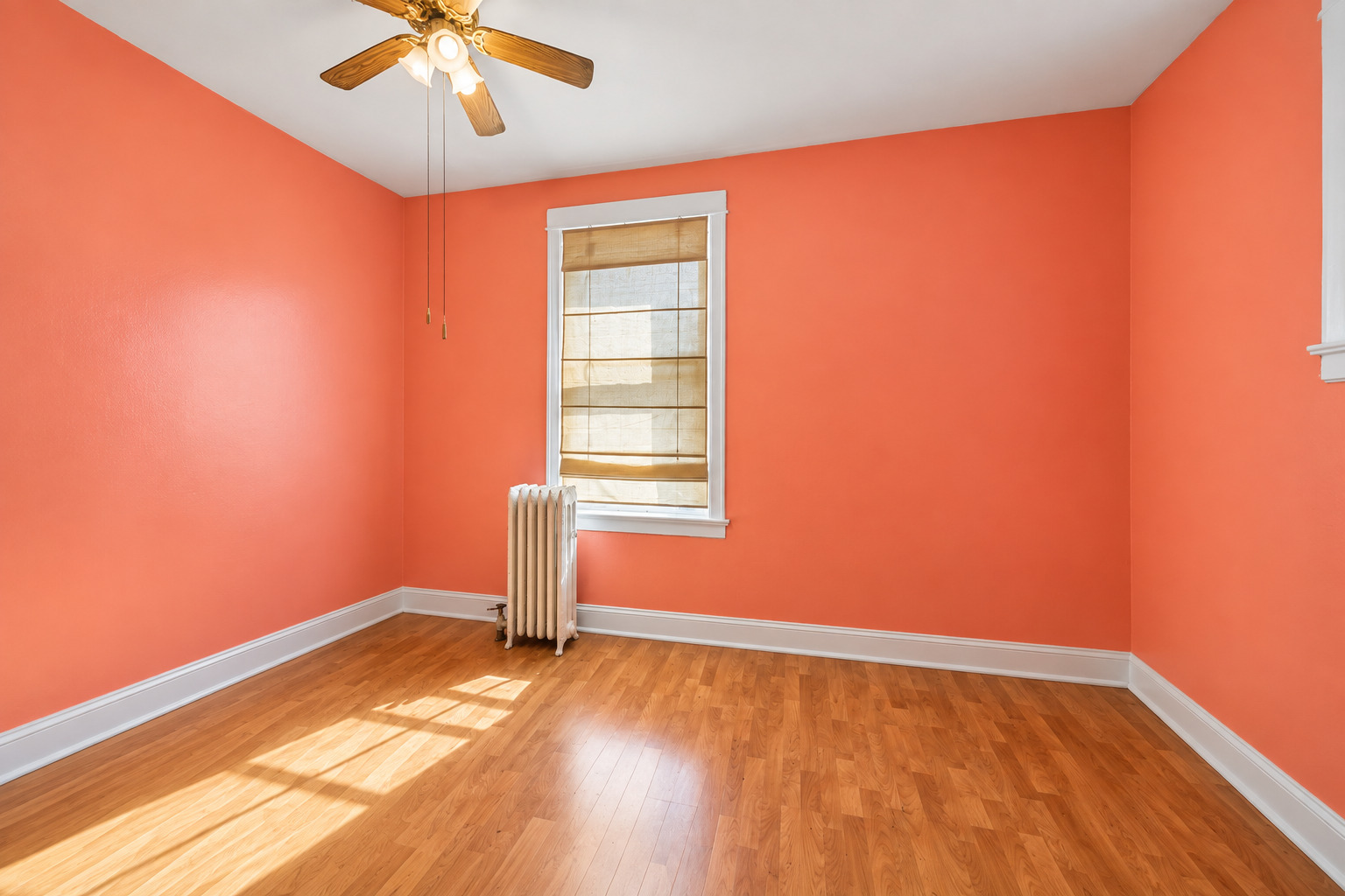 Empty bedroom with coral-orange walls, hardwood floors, and sunlight streaming through a shaded window.