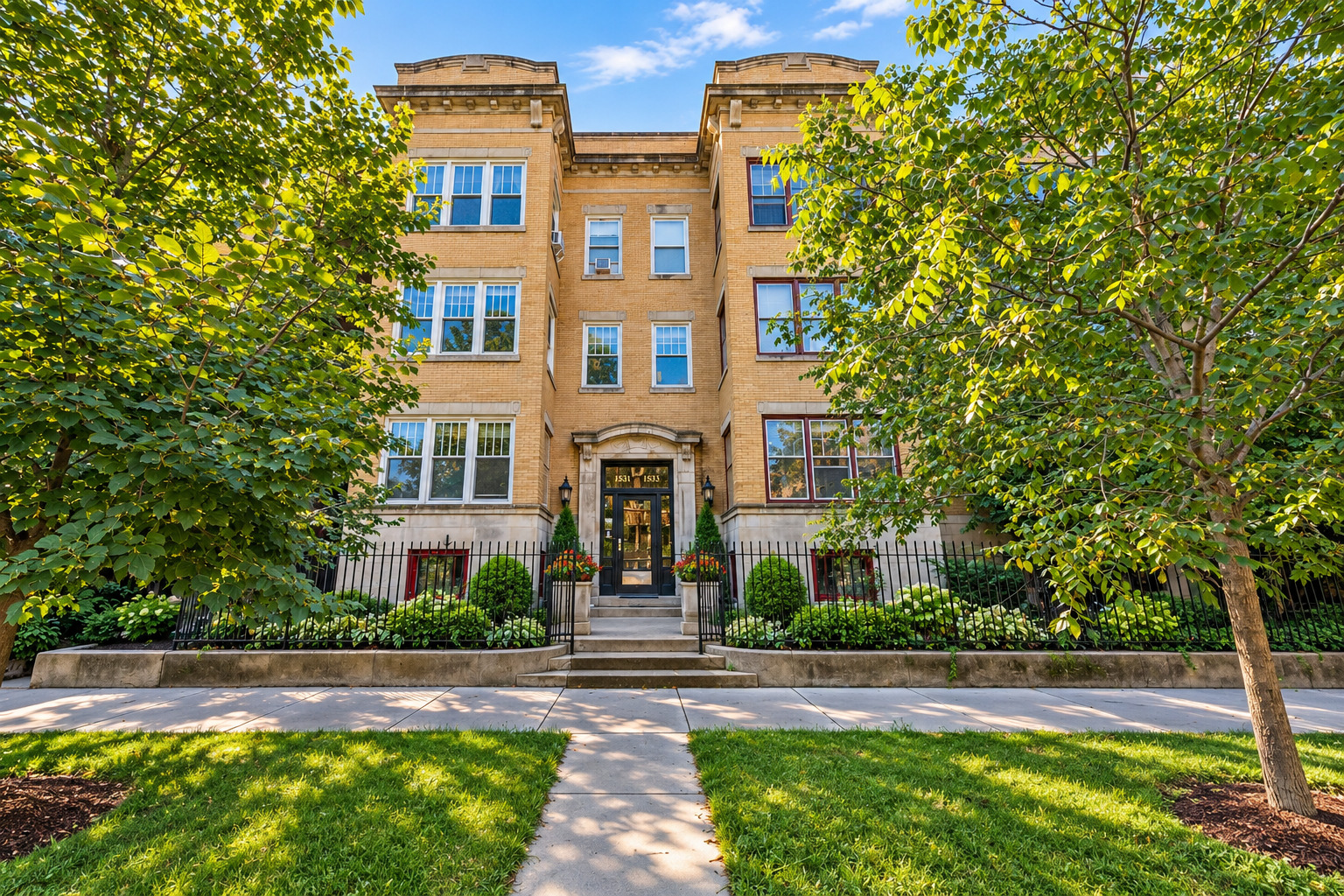 Tan brick apartment building with a central entrance, iron fence, and leafy trees.