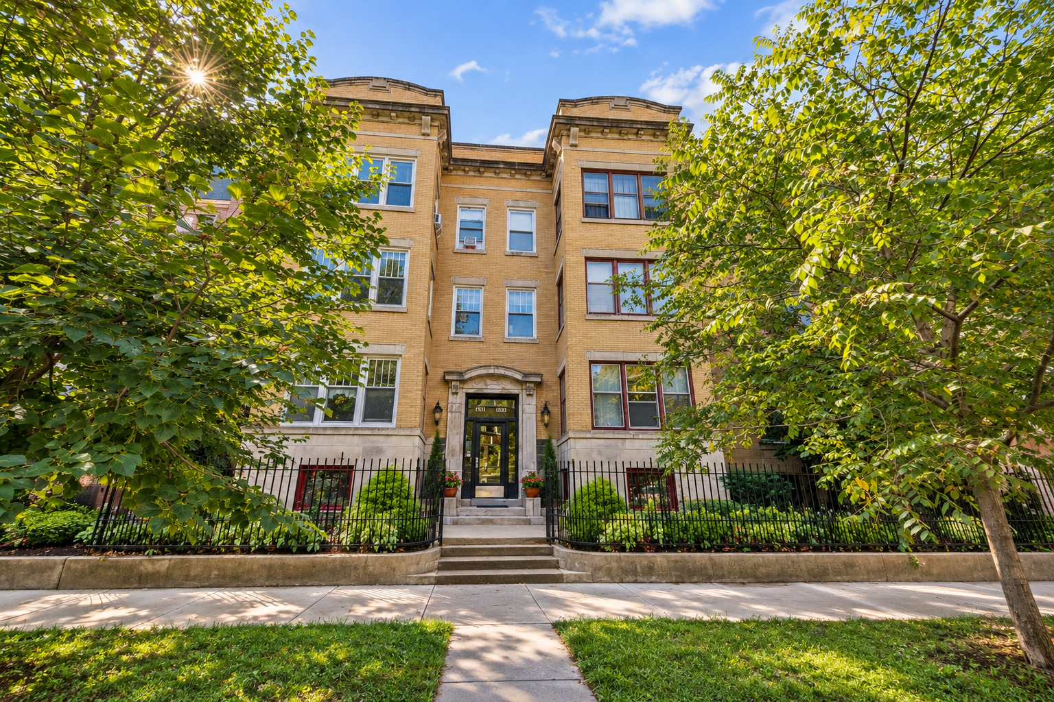 Tan brick apartment building with a central entrance, iron fence, and leafy trees.