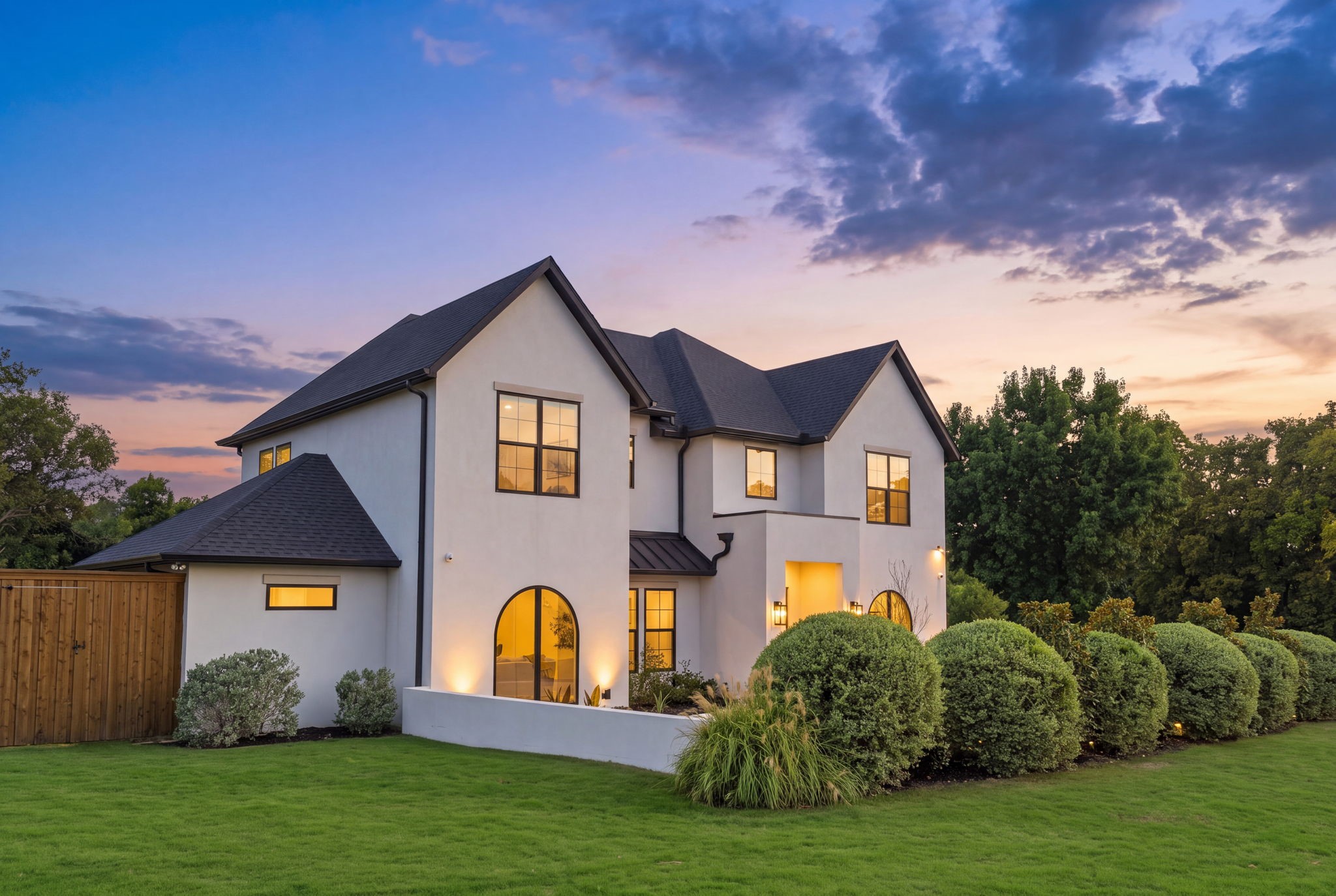 Modern two-story white house with glowing windows, an arched entry, and landscaped front lawn at dusk.