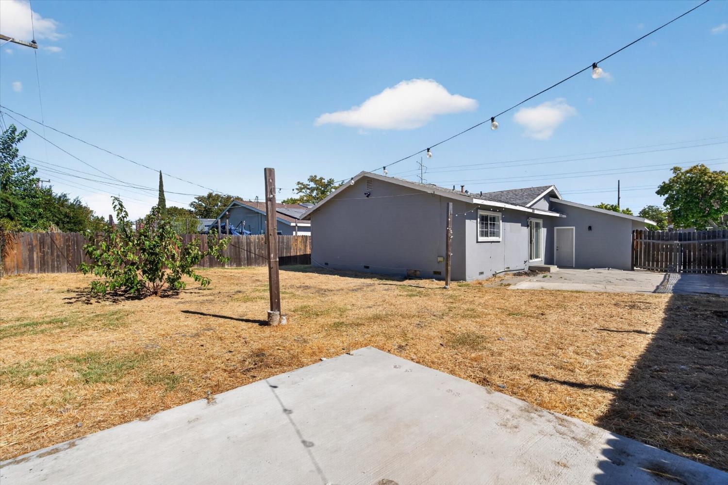 Gray home’s backyard with a concrete patio, dry lawn, wooden fences, and overhead string lights.