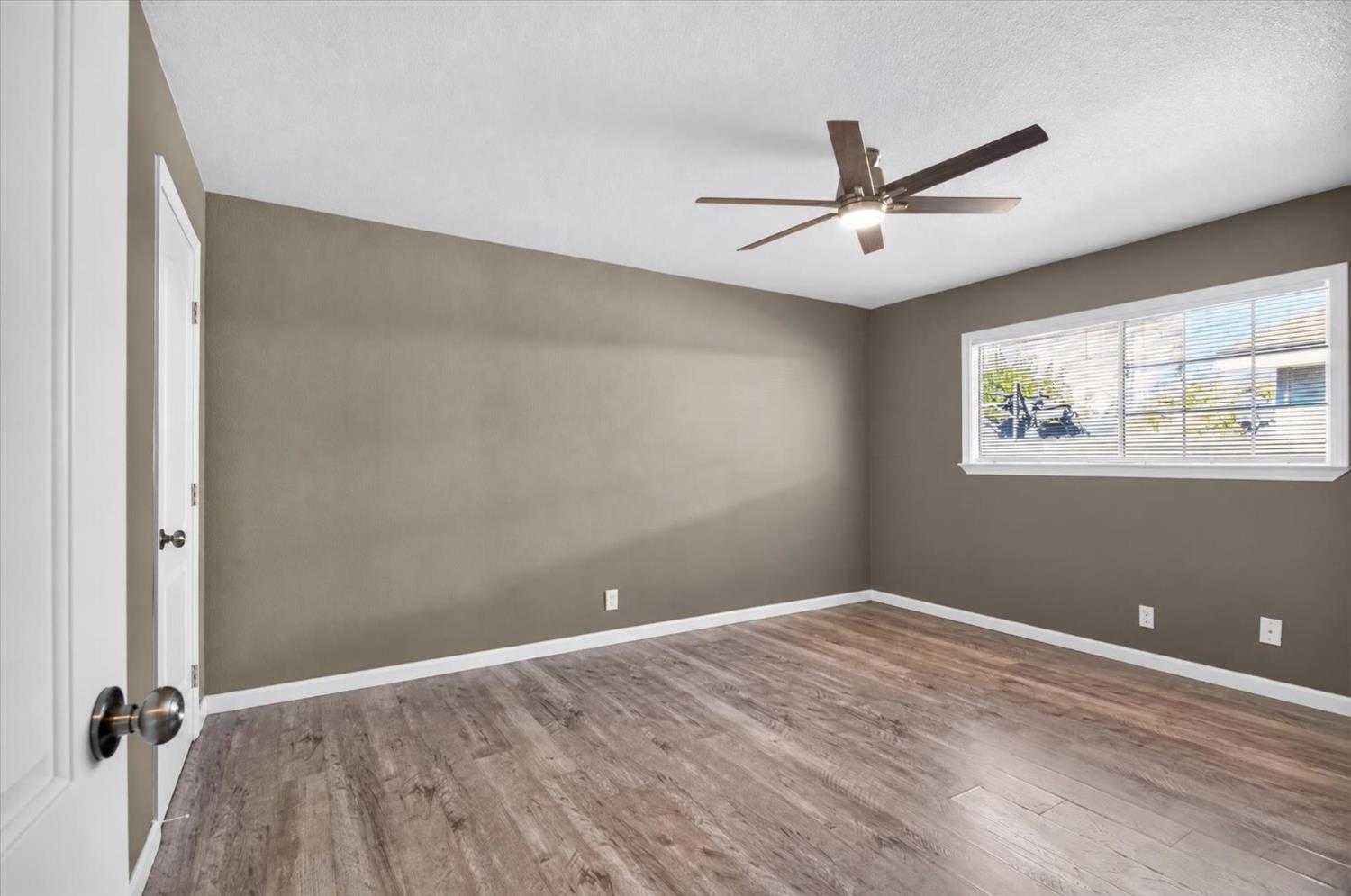 Empty room with wood-look flooring, a ceiling fan, and a wide window with blinds.