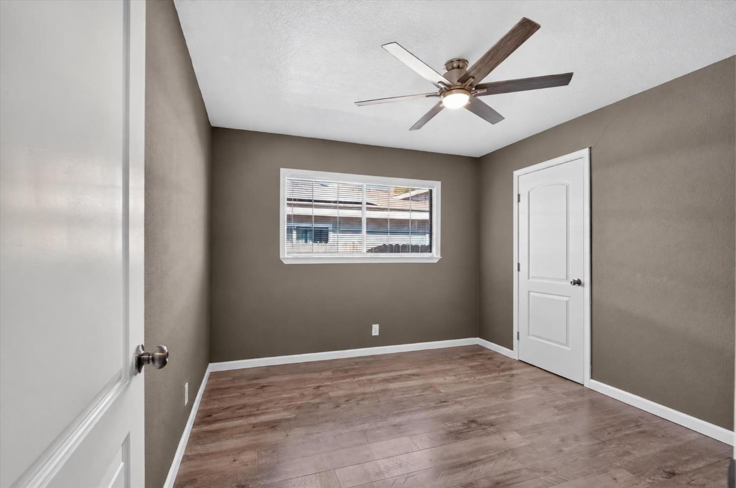 Empty bedroom with wood-look floors, taupe walls, a ceiling fan, and a window with blinds.