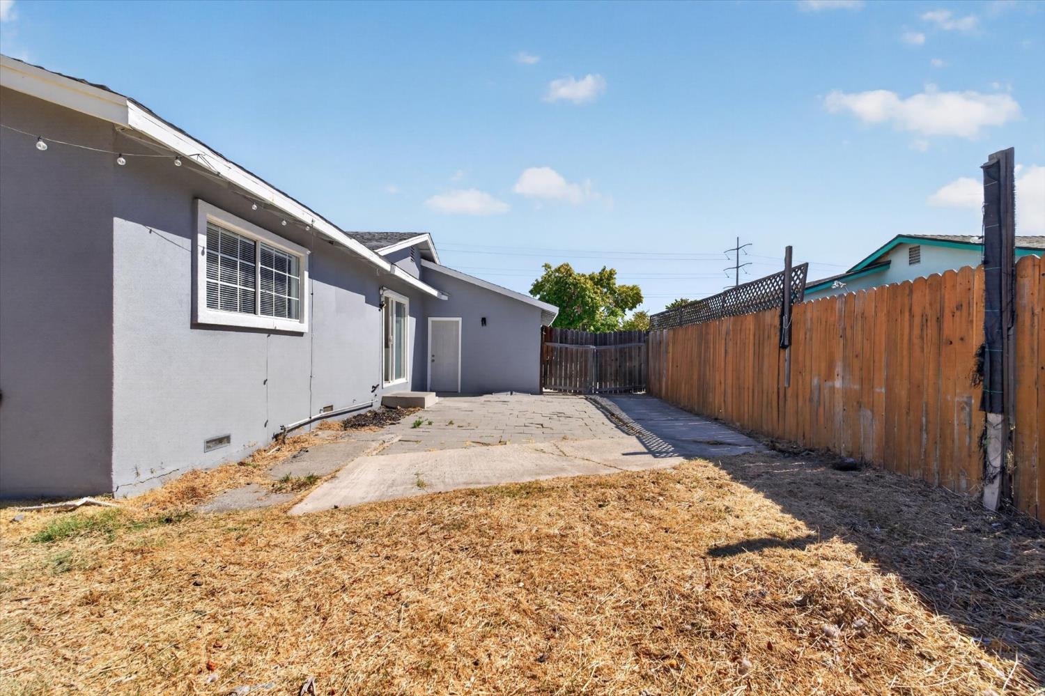 Backyard with a paved patio, dry lawn, and wooden privacy fence beside a gray single-story home.