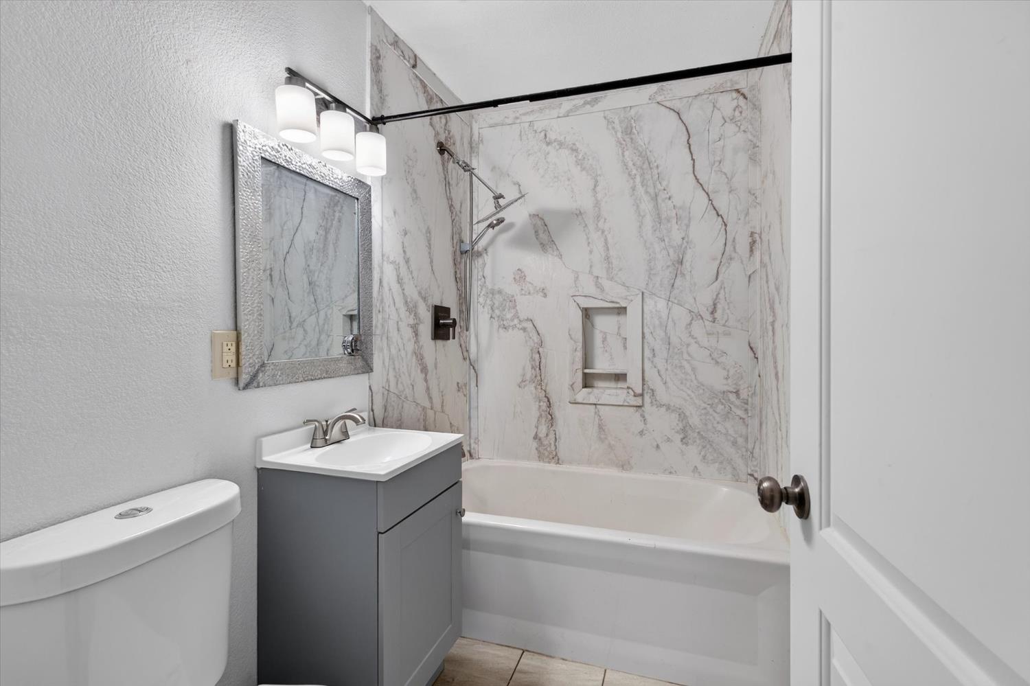 Bathroom with a marble-look tub and shower surround, recessed shelf, and gray vanity.