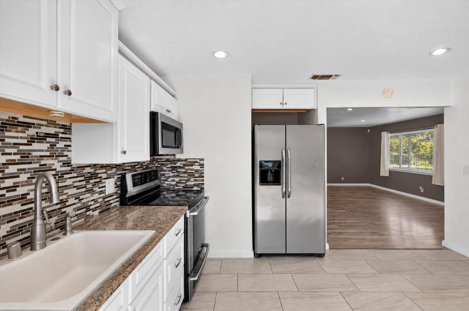 Kitchen with white cabinets, mosaic tile backsplash, granite counters, and stainless steel appliances opening to an unfurnished living room.