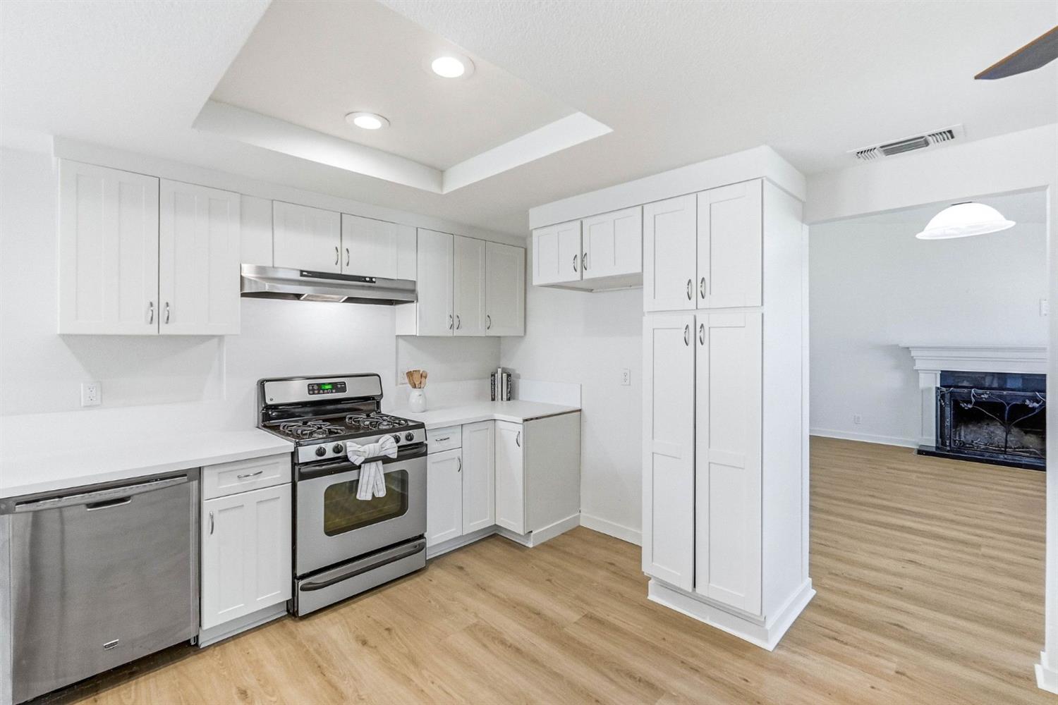 Kitchen with white cabinetry, stainless steel appliances, and light wood-look flooring beside a living room fireplace.