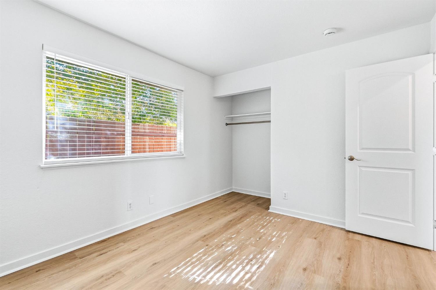 Empty bedroom with light wood floors, a wide window with blinds, and an open closet.