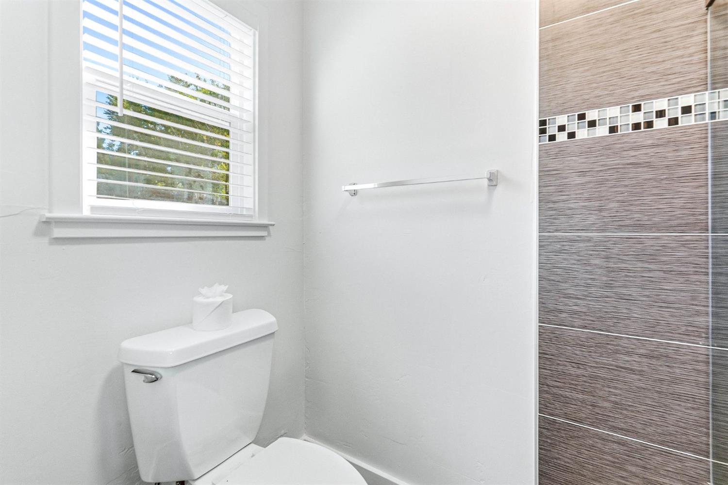 Bathroom with a white toilet, bright window, and gray-brown tiled shower wall.